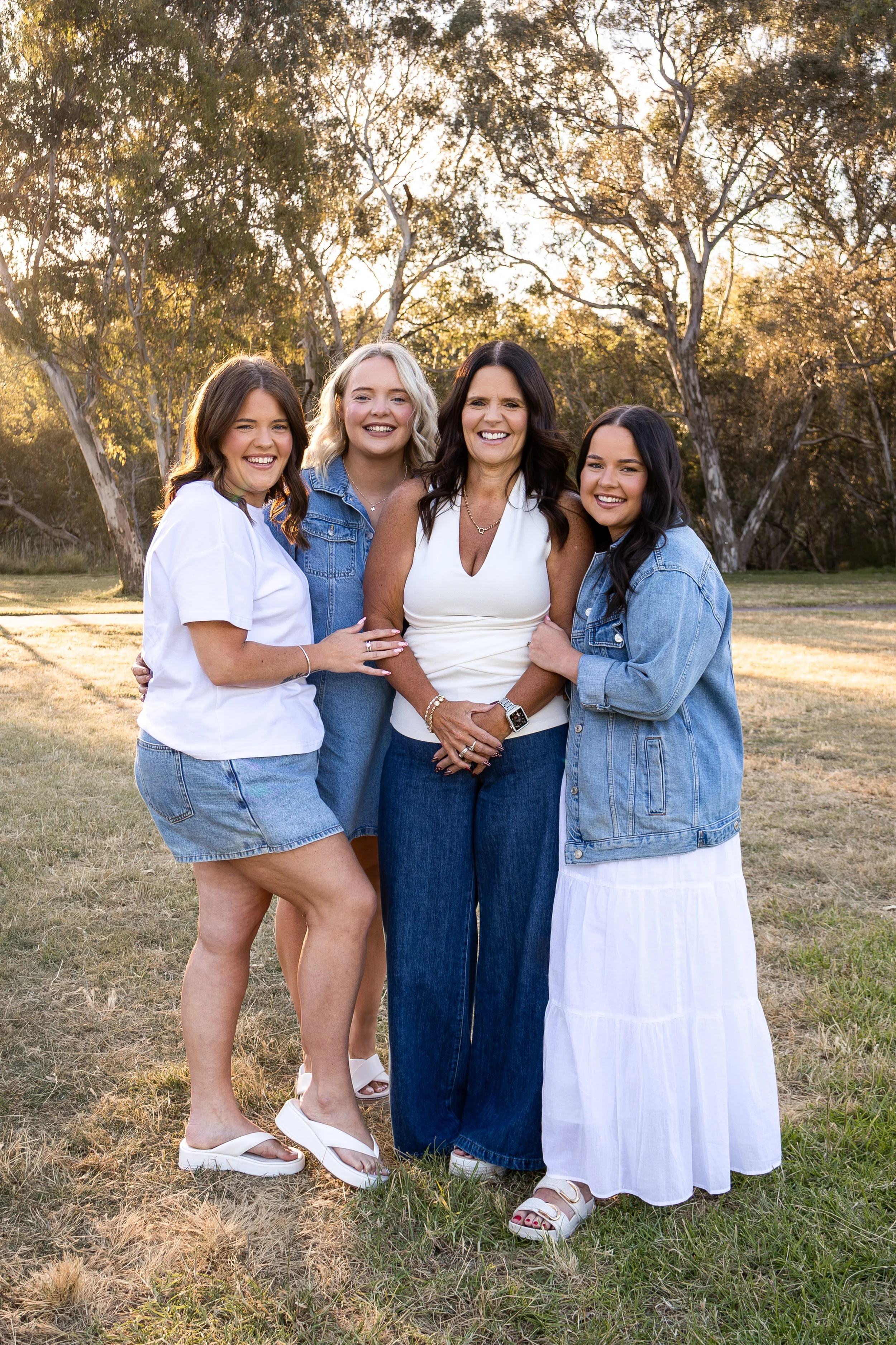 A group of four women standing outdoors in a park with trees and grass, enjoying the sunset, smiling at the camera. The woman in the center wears a white sleeveless top, and the others wear denim jackets or shirts and skirts or jeans.