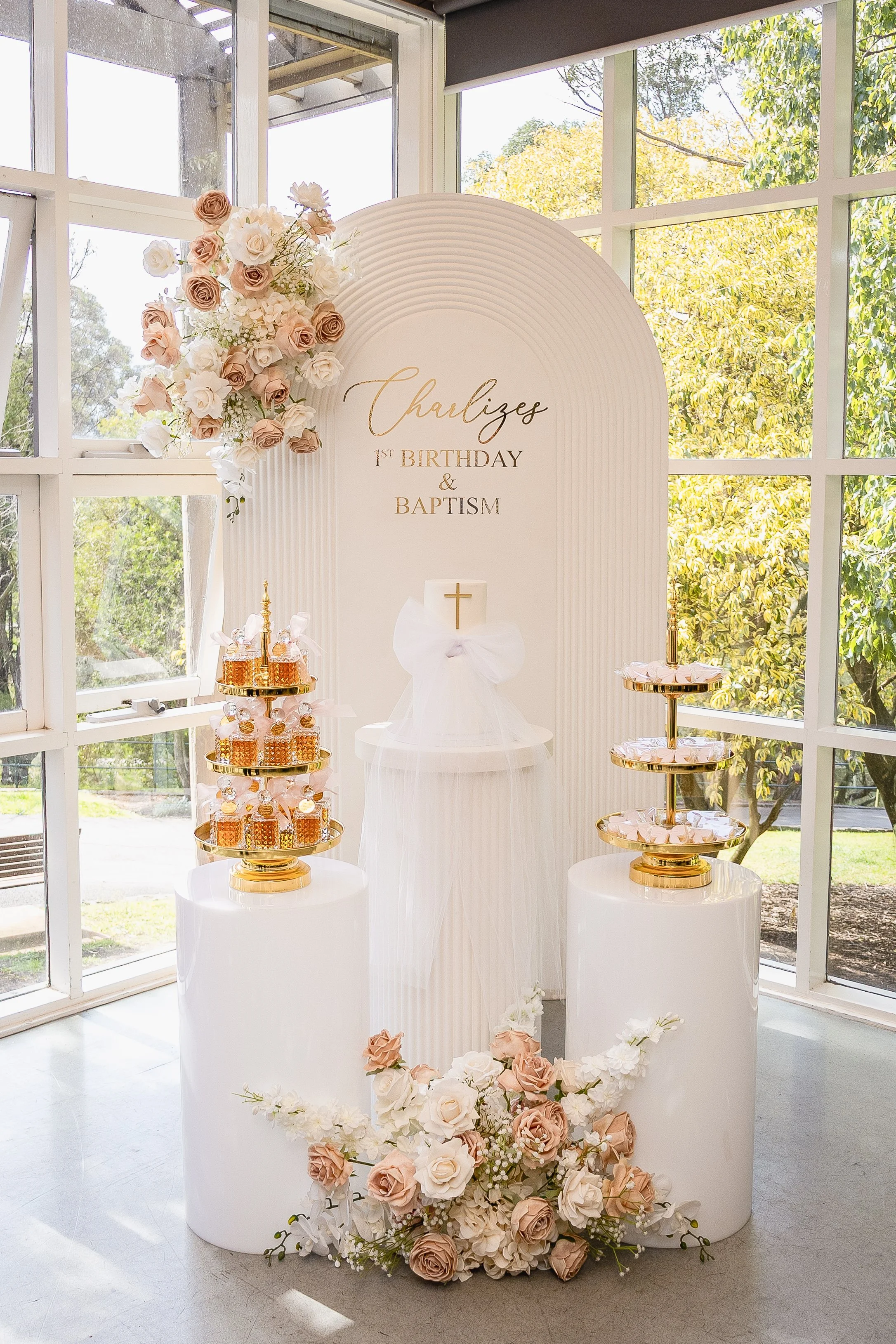 Decorative display for a child's first birthday and baptism, featuring a floral arrangement, a cake with a cross, and sweet treats on gold trays, set in a bright room with large windows.