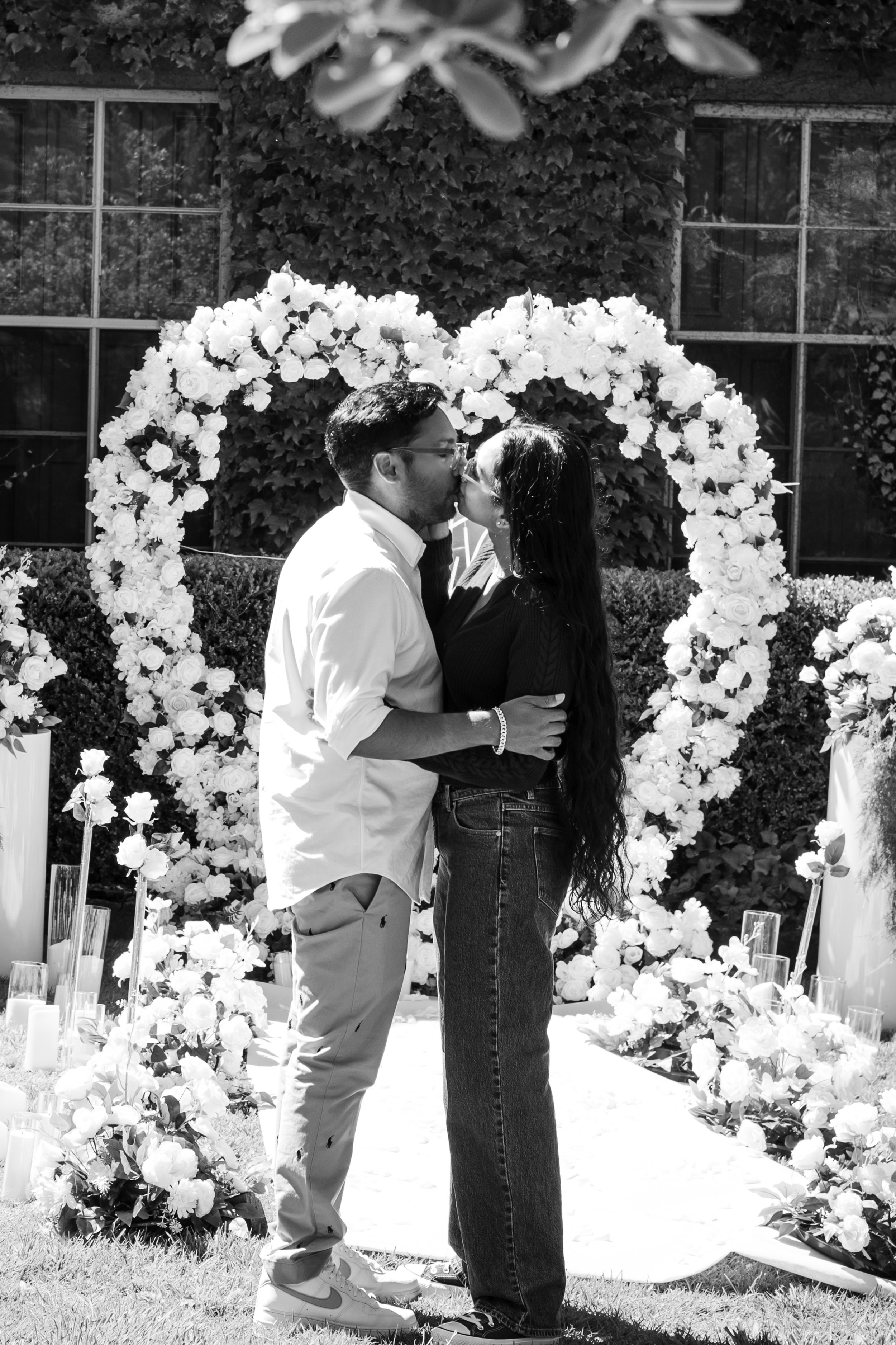A couple kissing in front of a floral heart-shaped arch during a wedding proposal outdoors in black and white.