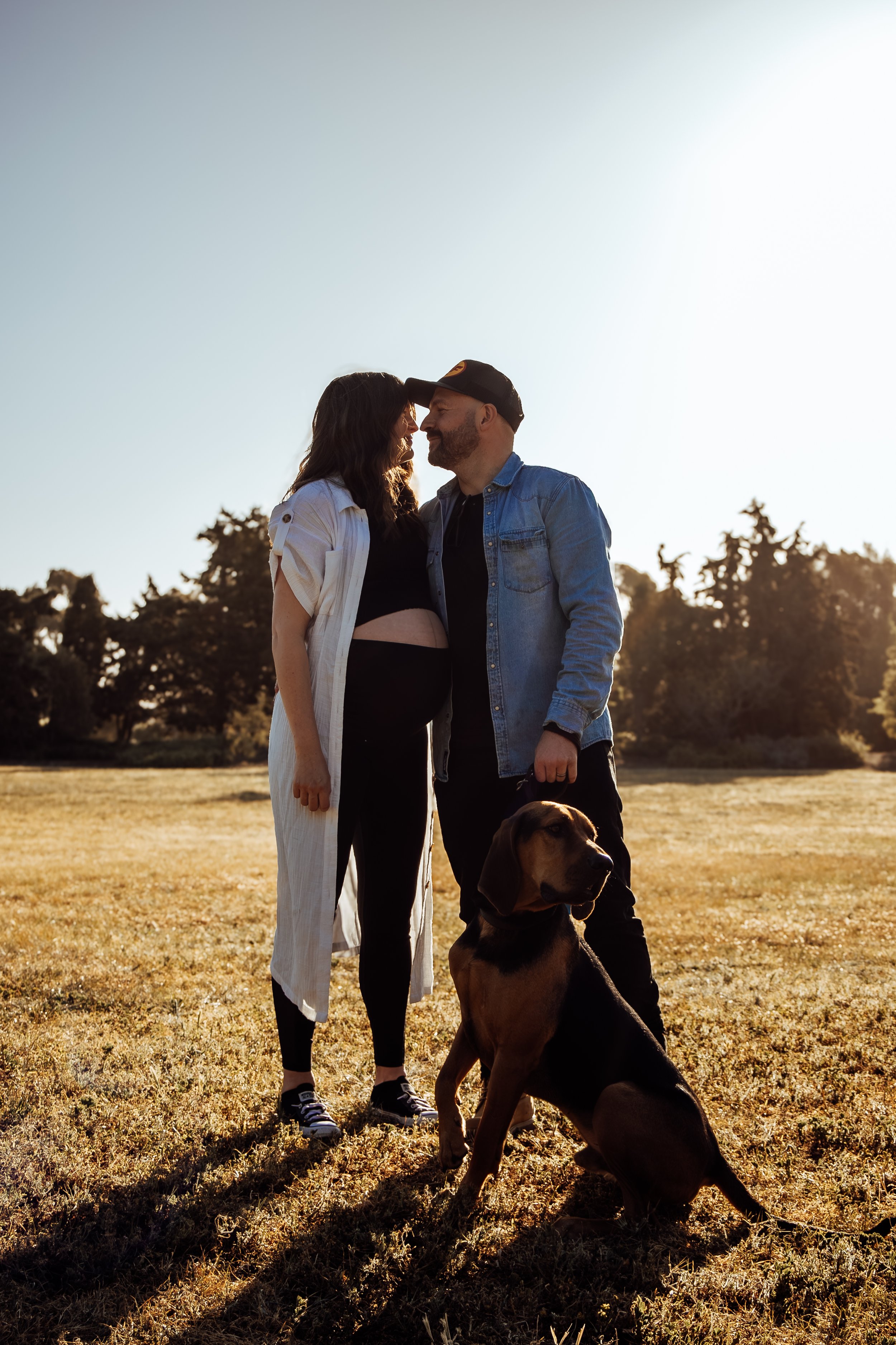 A couple and their dog outdoors during sunset, standing close with foreheads touching, smiling, with trees in the background.