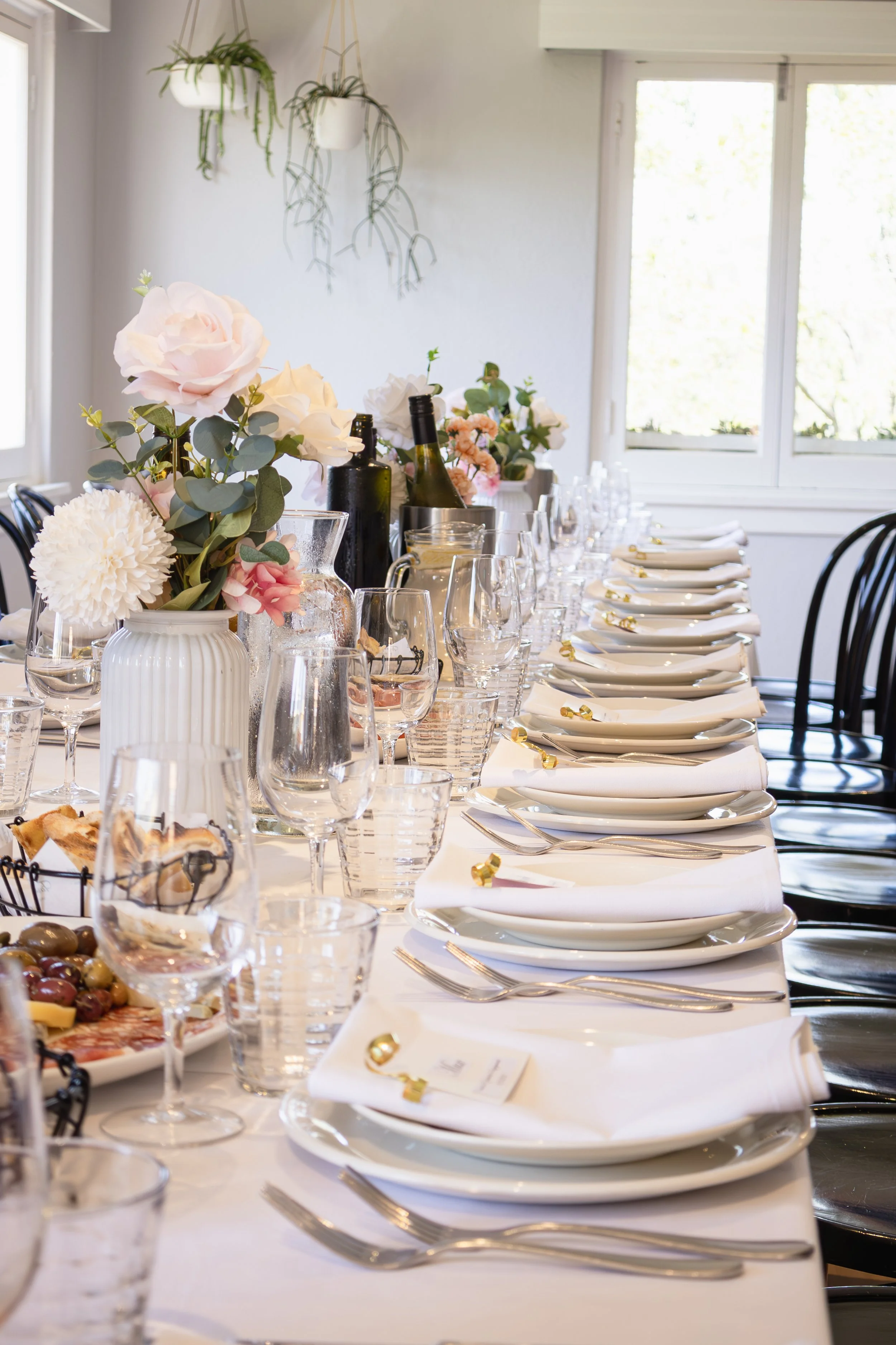 A long dining table set for a formal event with white plates, napkins, silverware, wine glasses, and wine bottles. The table is decorated with pink and white flower arrangements in vases. Sunlight streams through the windows in a bright, modern room.