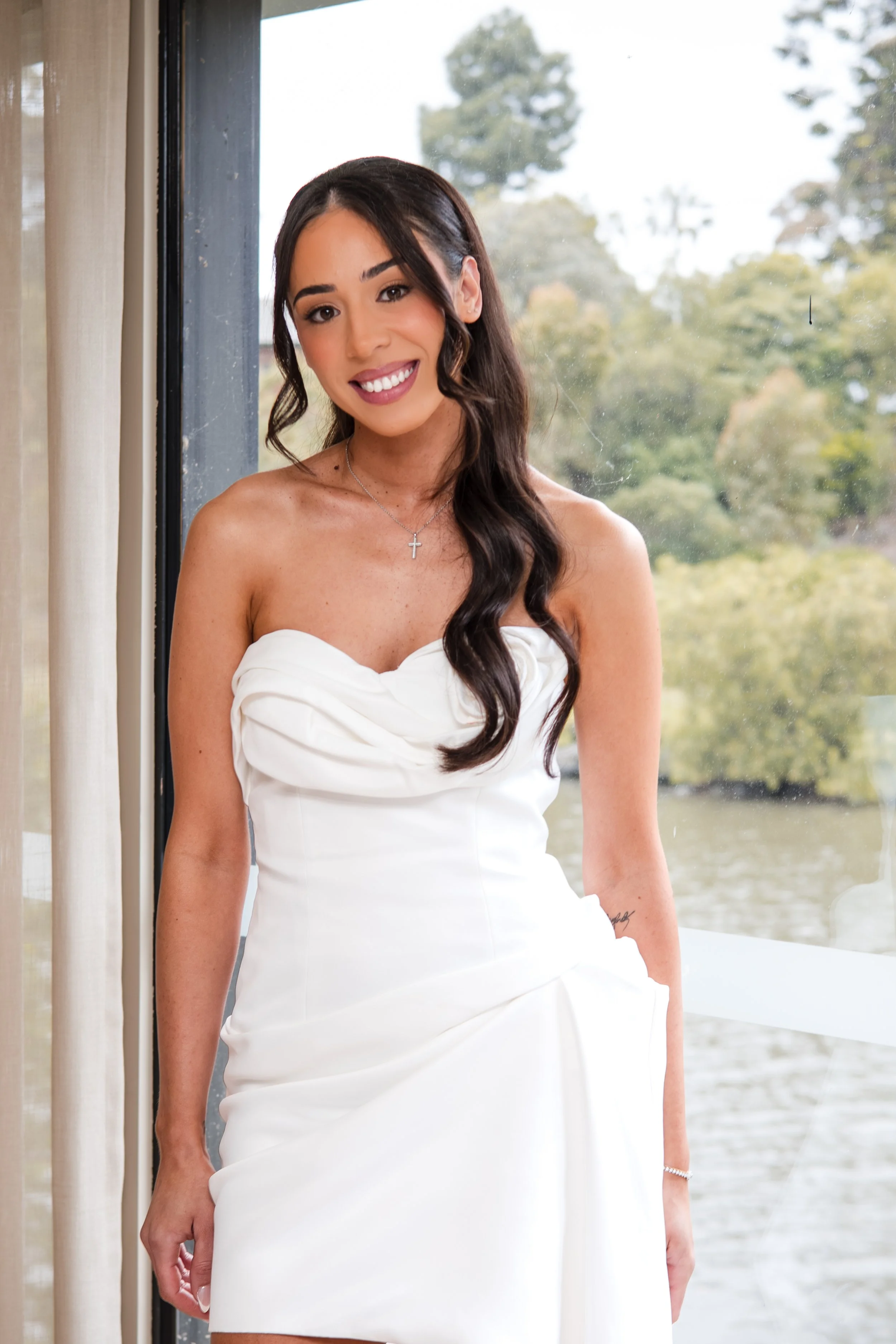 A young woman in a white strapless dress posing by a window with a lake and trees in the background.