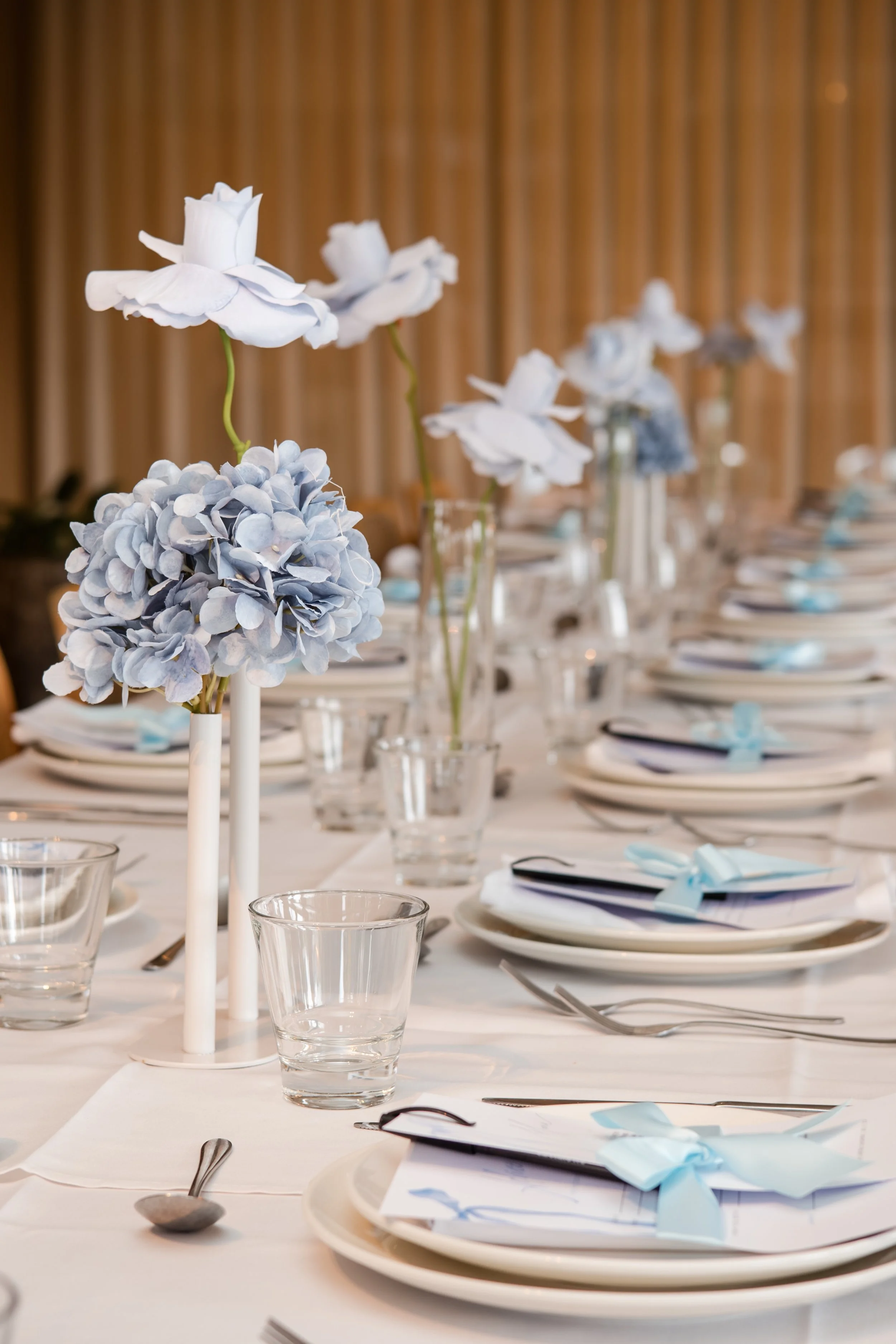 Elegant dining table set with white plates, silverware, clear glasses, and light blue ribbon-tied napkins. Tall flower arrangements with white and light purple flowers serve as centerpieces, on a table with a white tablecloth.