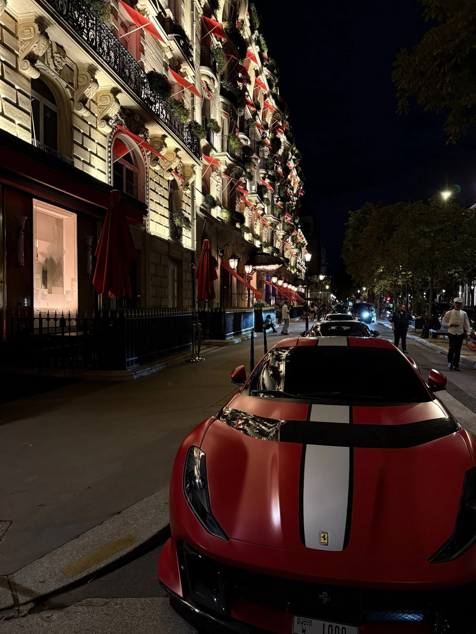Night scene on a city street featuring a red Ferrari sports car with black and white racing stripes in the foreground, a multi-story building with ornate architectural details and red awnings, some outdoor seating with closed umbrellas, pedestrians walking along the sidewalk, and trees with streetlights illuminating the area.