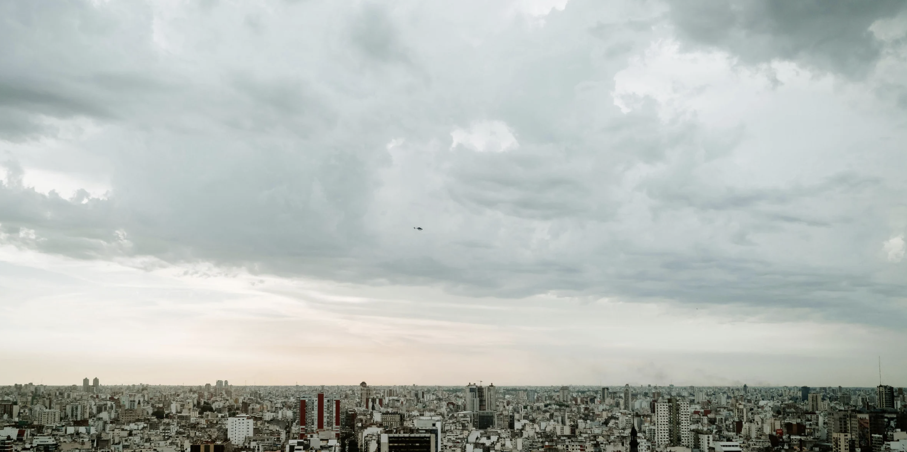 Overcast cityscape with numerous buildings under a cloudy sky, with a small helicopter flying in the distance.