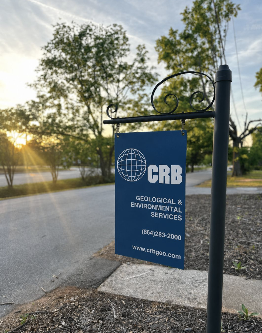 Sign for CRB Geology & Environmental Services hanging on a black metal post outdoors during sunset. Trees and a road are in the background.