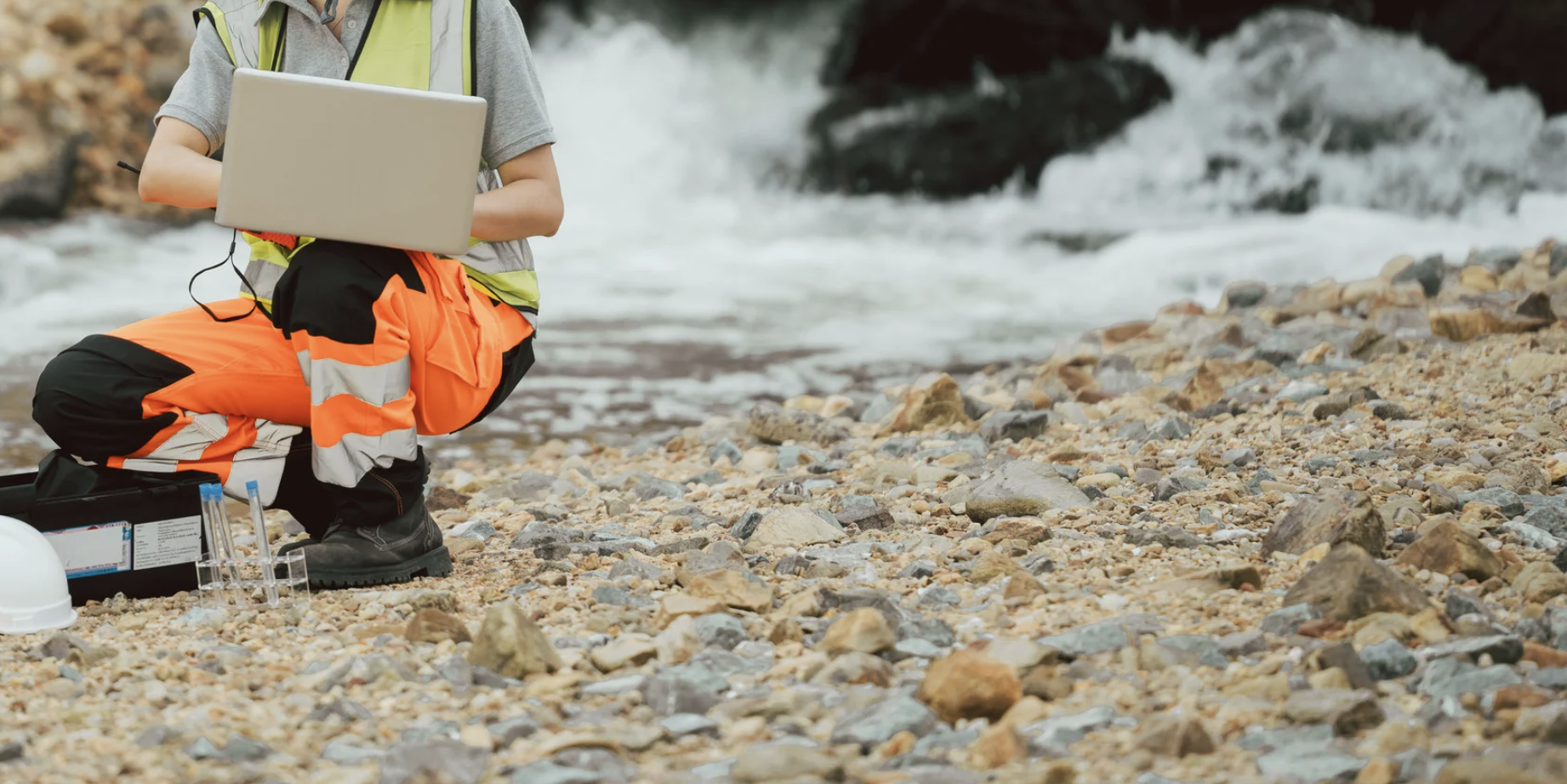 A person in high-visibility orange and black protective clothing kneeling on a rocky beach near the water, working with a laptop and scientific equipment.