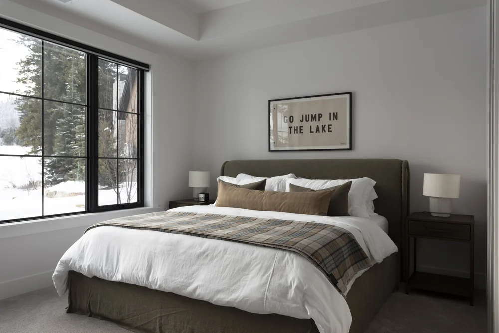 Minimalist bedroom with olive upholstered bed, plaid throw, and snowy evergreen view through black-framed window