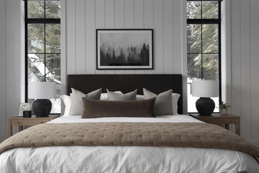 Minimalist bedroom with olive upholstered bed, plaid throw, and snowy evergreen view through black-framed window