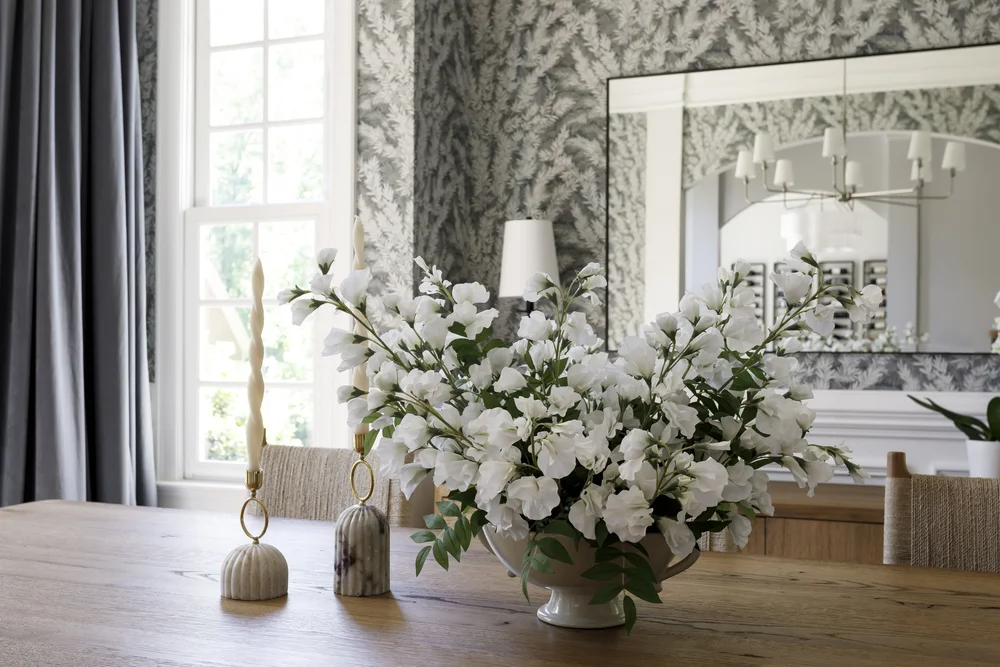 Dining table with white flower bouquet before fern-patterned wallpaper, a large mirror, and window