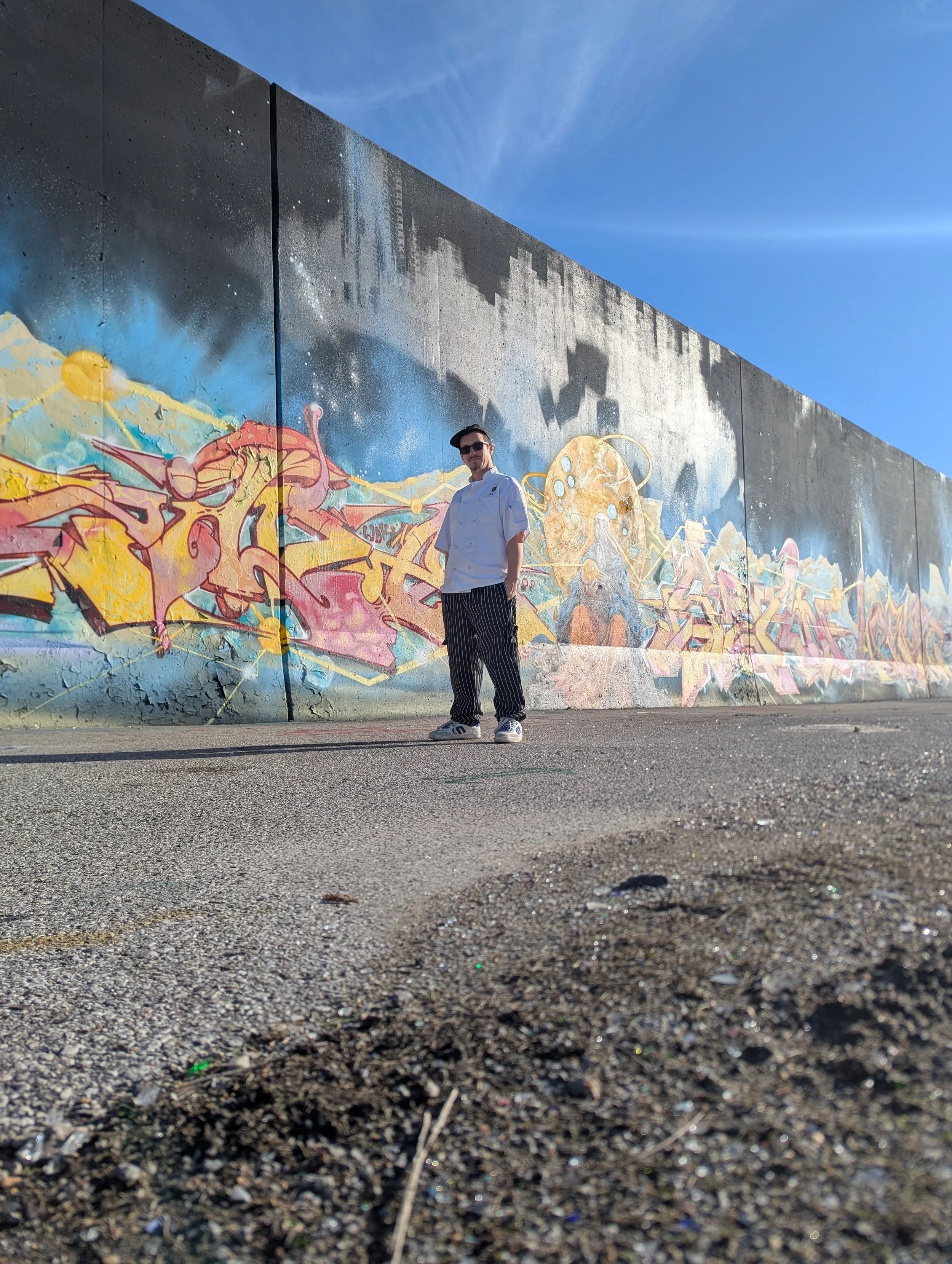 A man in a white chef's coat and black striped pants standing in front of a colorful graffiti mural on a concrete wall under a blue sky.