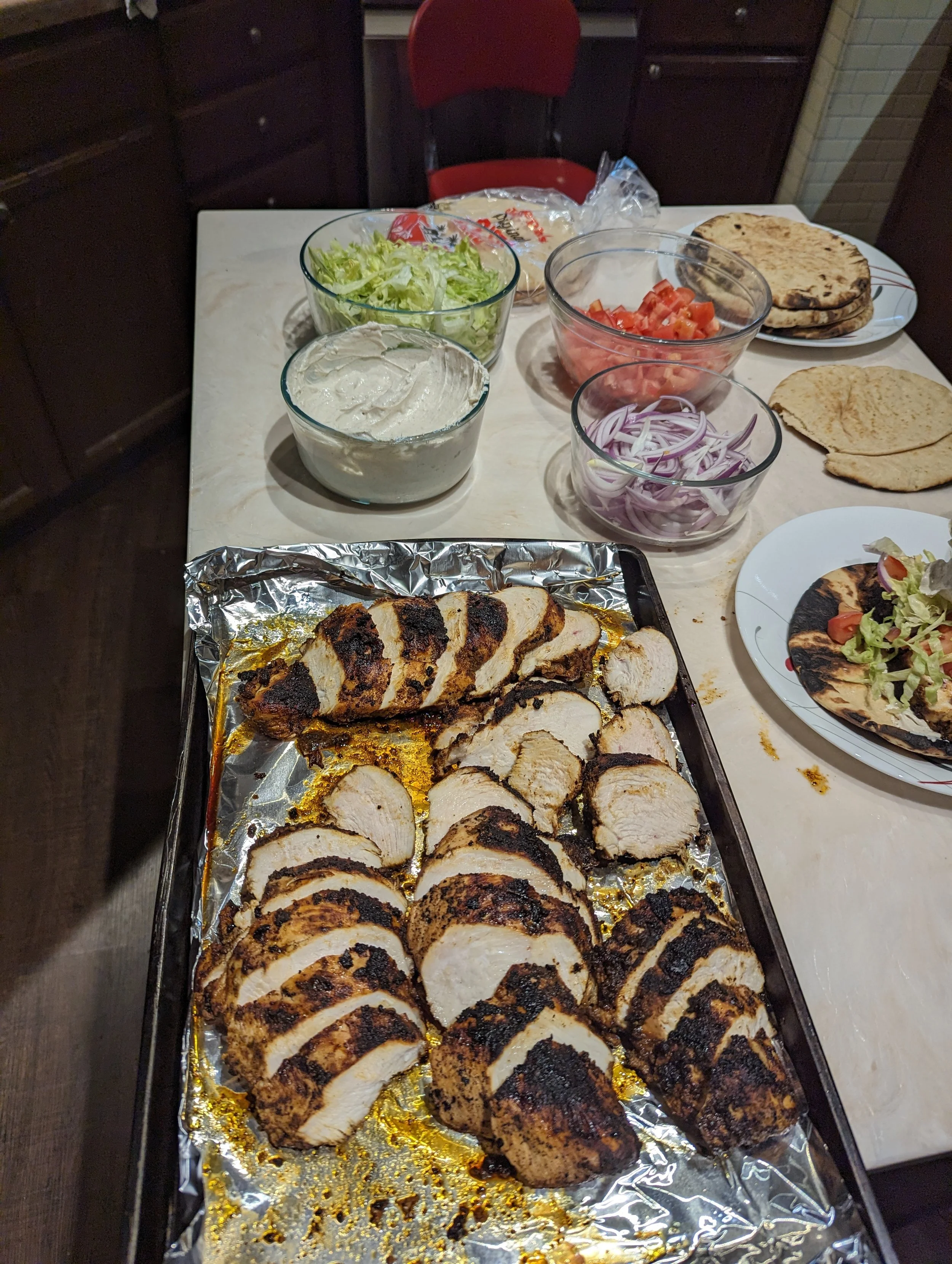 Sliced baked chicken breast with herbs on a baking sheet, surrounded by bowls of lettuce, tomatoes, red onions, hummus, pita bread, and a plate with salad and grilled pita.