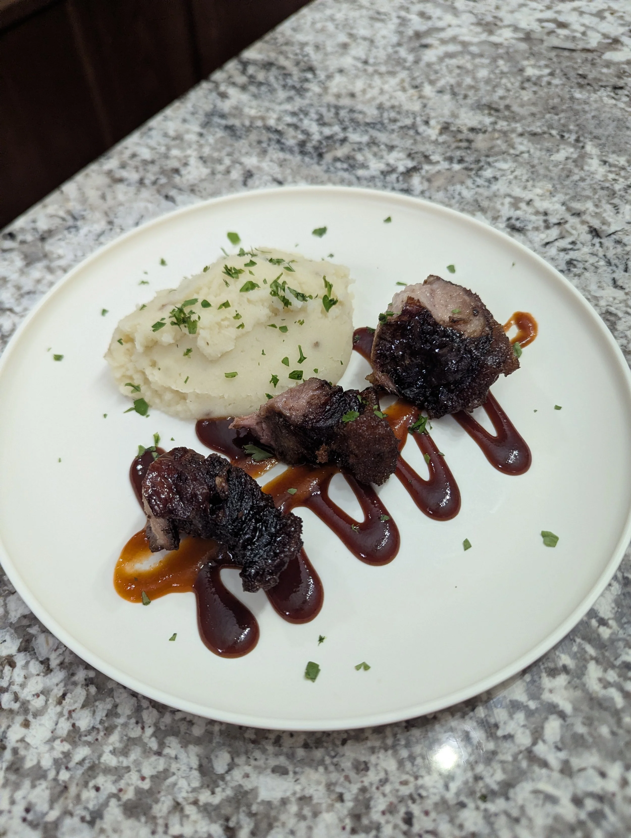 Plate of mashed potatoes and three pieces of grilled meat with sauce, garnished with chopped herbs.