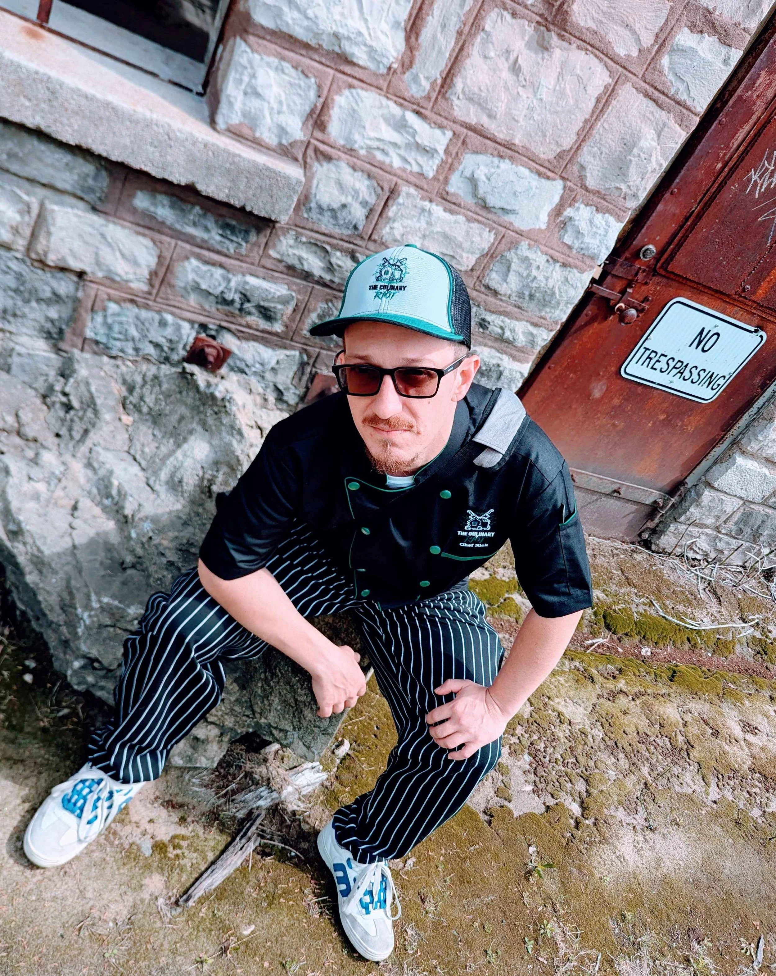 A Chef in Valmeyer, IL wearing glasses, a baseball cap, a chef's jacket, and striped pants, crouching outdoors on mossy ground near a stone wall, with a 'No Trespassing' sign on a wooden structure nearby.