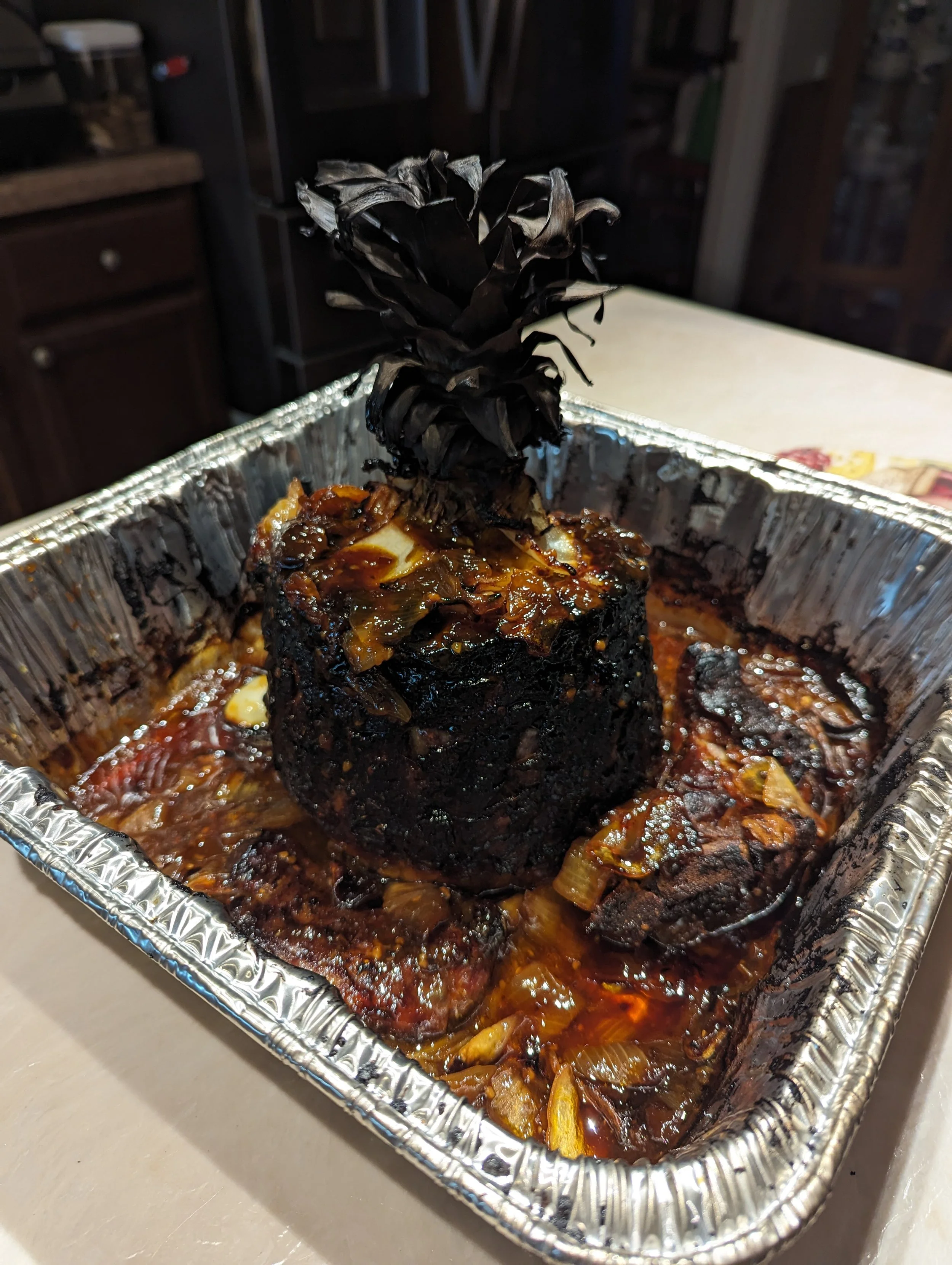 A baked dish in a foil container featuring a small pineapple with darkened, caramelized exterior, surrounded by sauce and cooked vegetables.