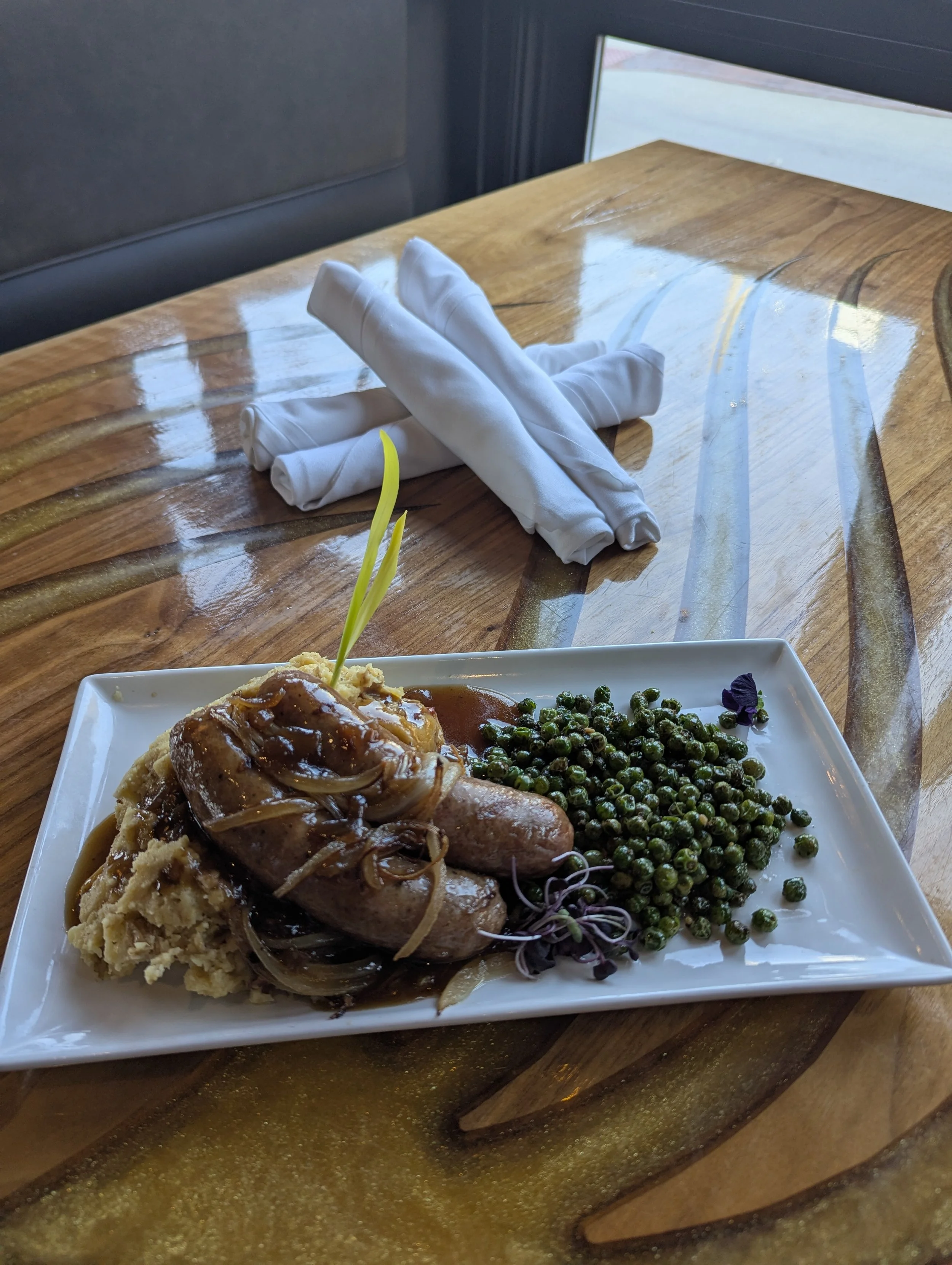 Plate of mashed potatoes with gravy, sausage links, cooked green peas, and garnishes on a wooden table, with neatly folded white napkins in the background.