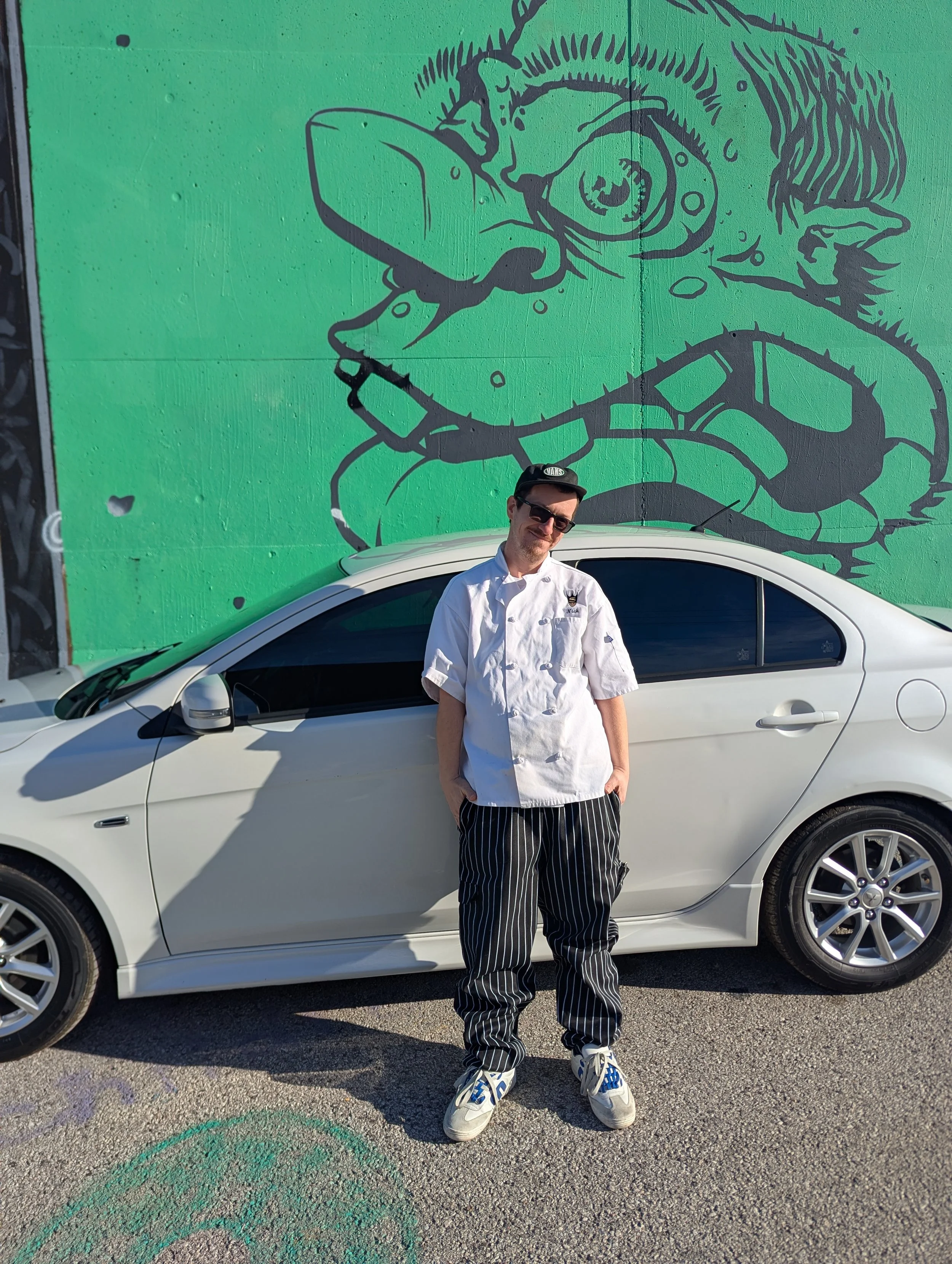 Man in chef's coat and striped pants standing in front of a white car, with a green mural mural featuring a large, stylized drawing of an animal wearing glasses behind him.