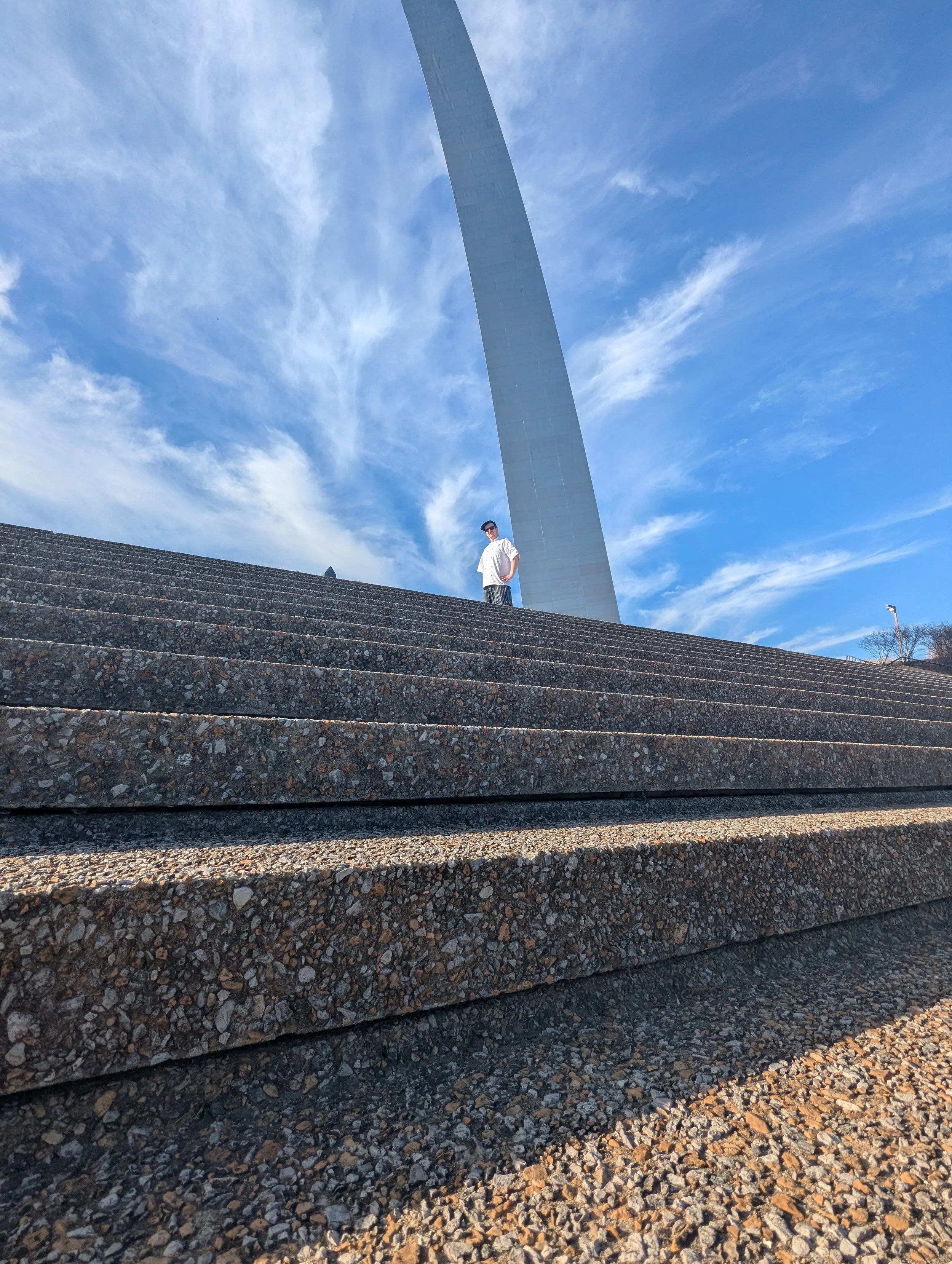 Person standing on concrete steps with the Gateway Arch in St. Louis, Missouri in the background, clear blue sky and wispy clouds.