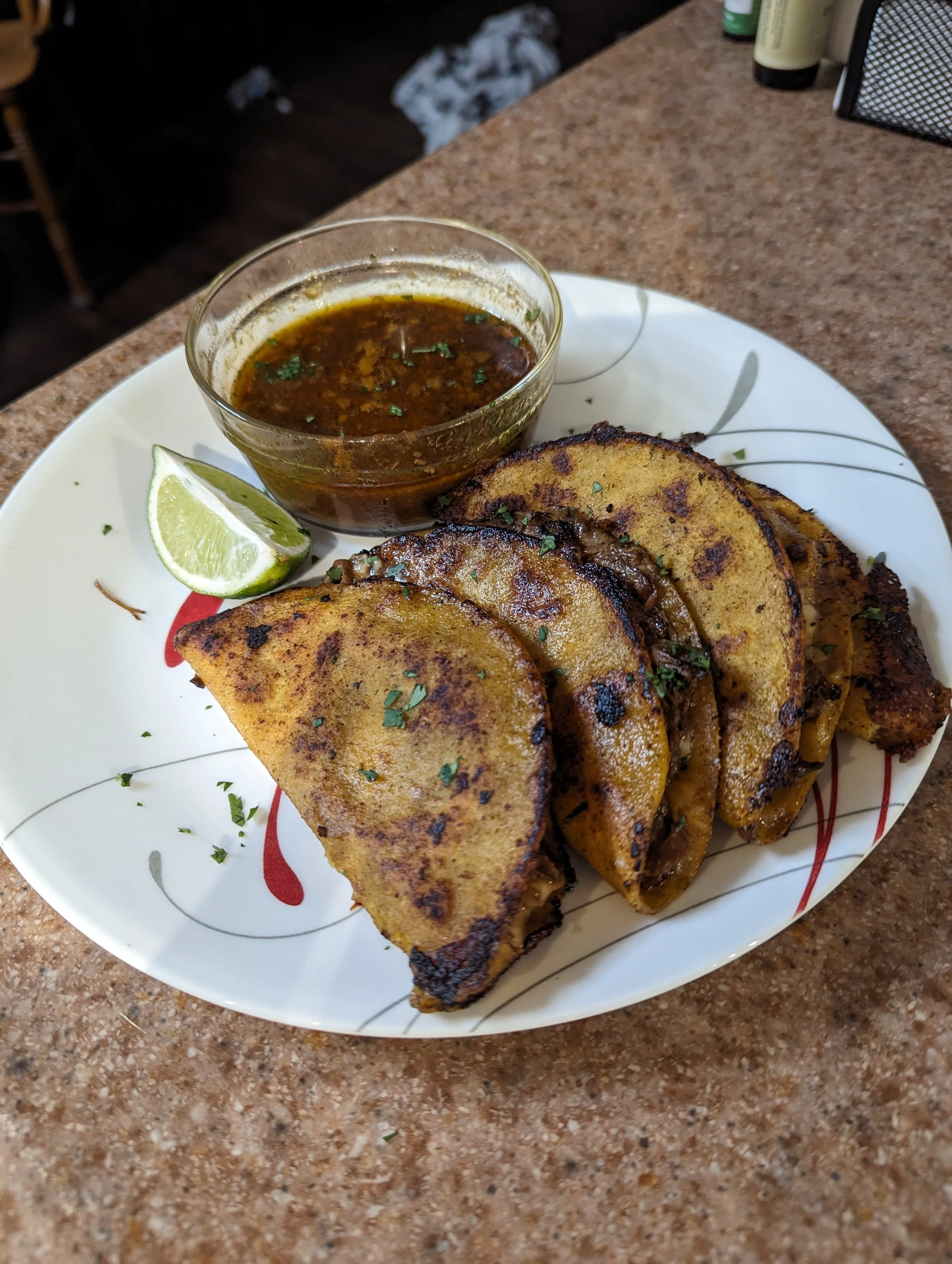 Four grilled steak tacos with chopped cilantro on a white plate, lime wedge, and a bowl of dark sauce on a brown countertop.