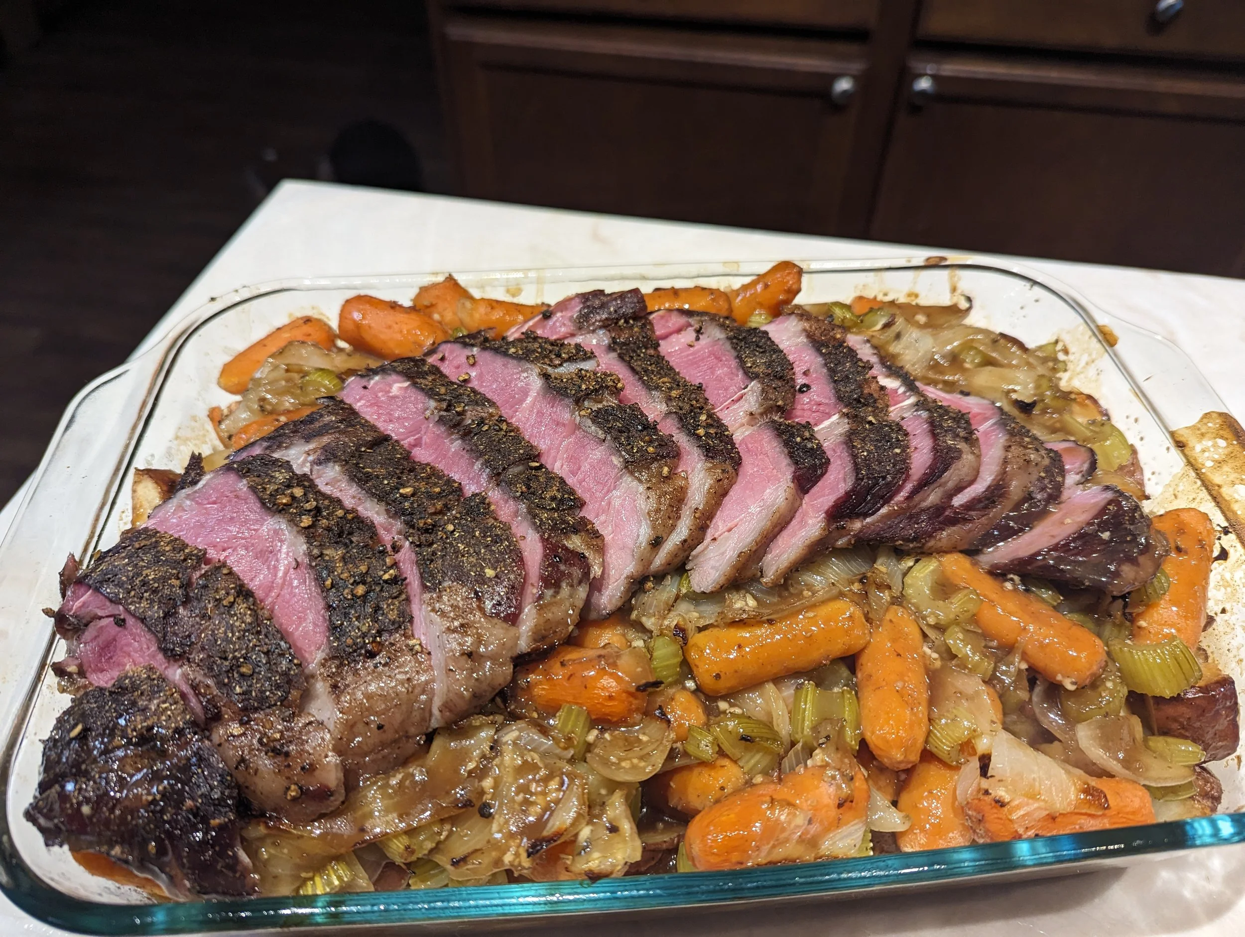 Baked dish with sliced peppercorn-crusted steak on top of roasted vegetables including carrots, onions, and celery in a glass baking dish.