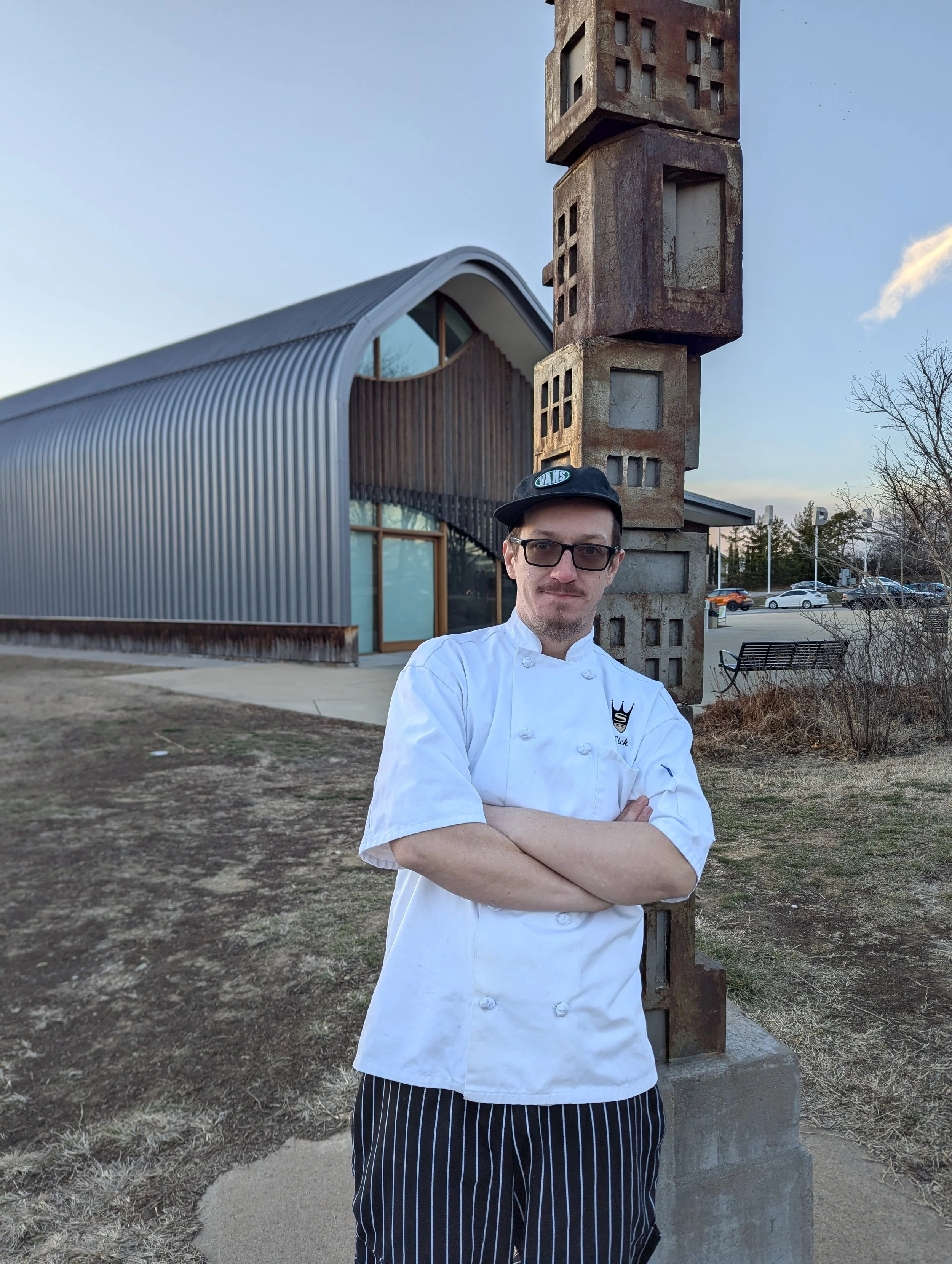 A man in a white chef uniform and black hat with glasses standing outdoors with arms crossed, next to a large sculpture made of concrete blocks, in front of a modern building with curved metallic siding.