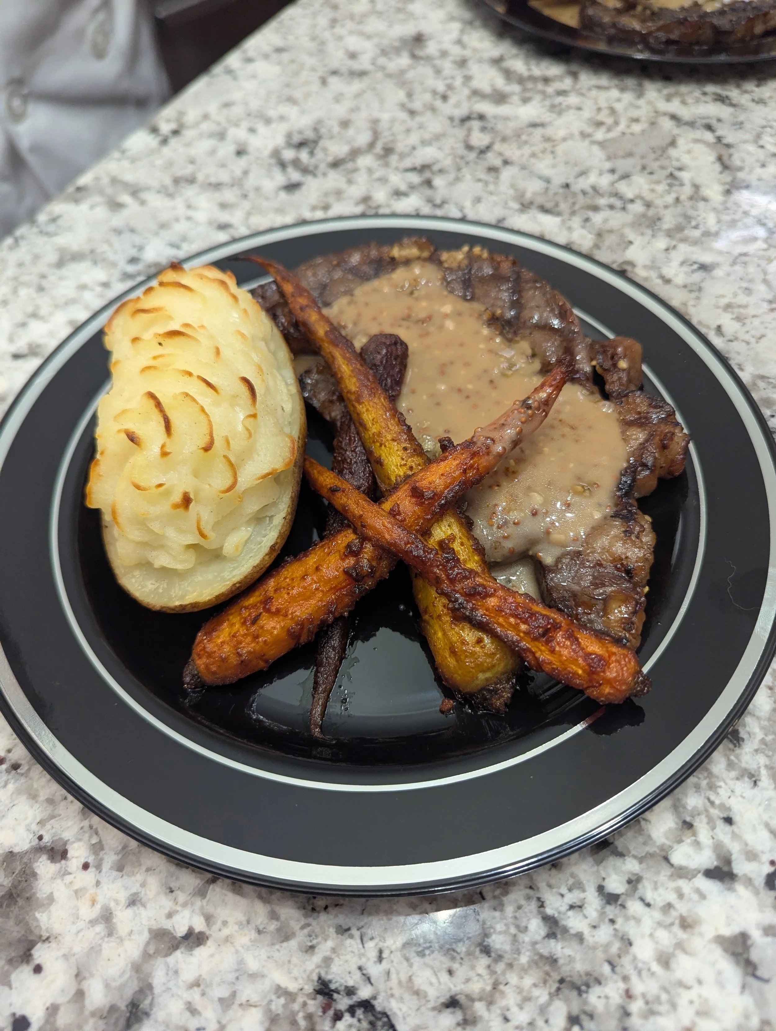 A plate of food with mashed potatoes, roasted carrots, steak with gravy, served on a black plate with a white rim on a granite countertop.