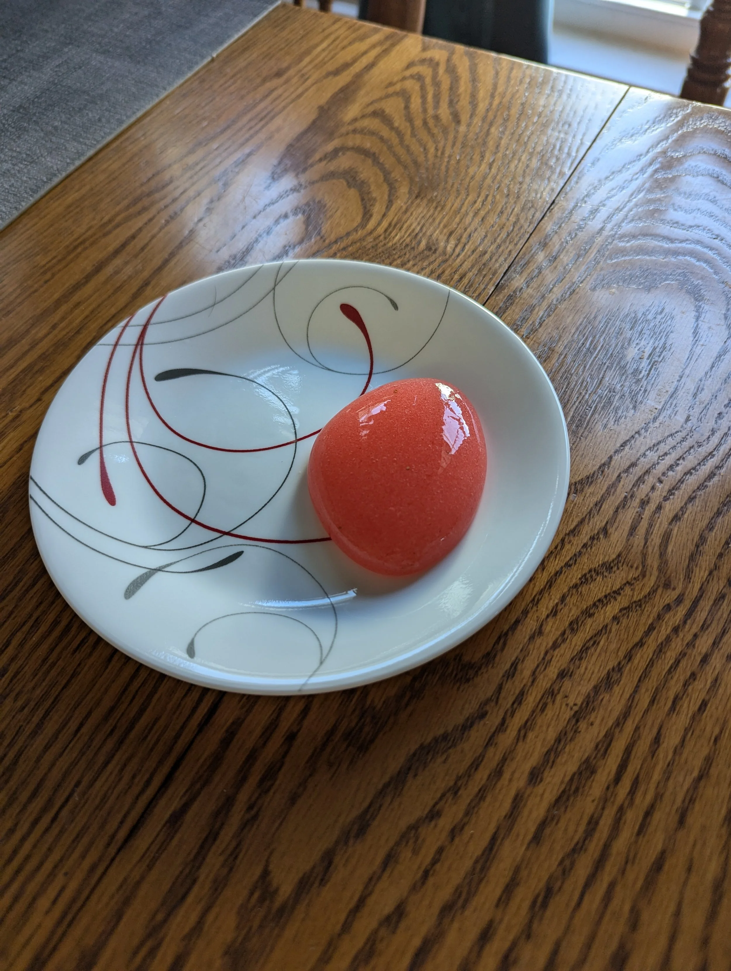 A red egg-shaped dessert on a white plate with colorful swirling line patterns, placed on a wooden table.