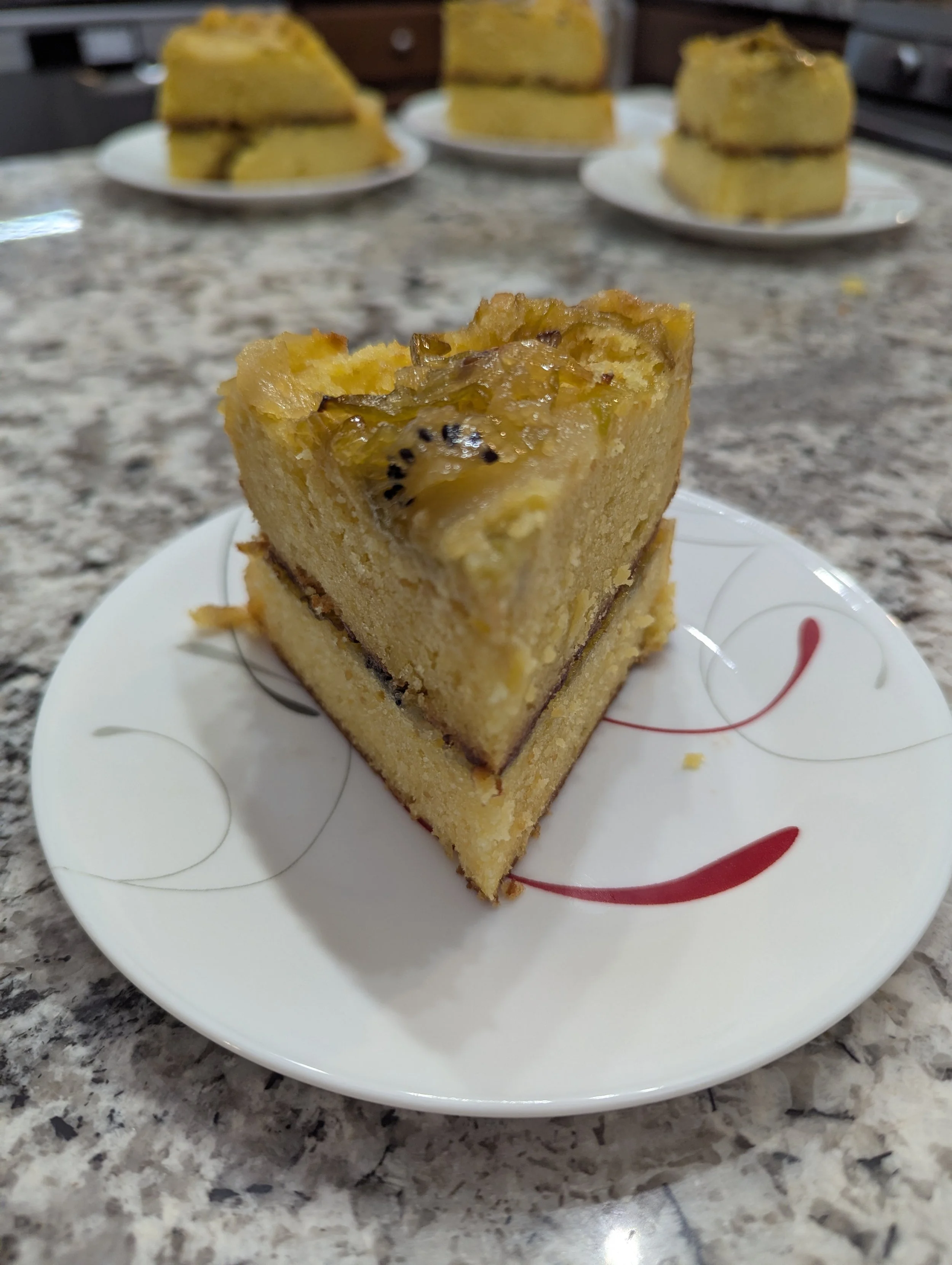 A slice of fruit cake with kiwi slices on top, on a white plate with red and black swirl design, on a granite countertop. In the background, three more slices of the same cake are on white plates.