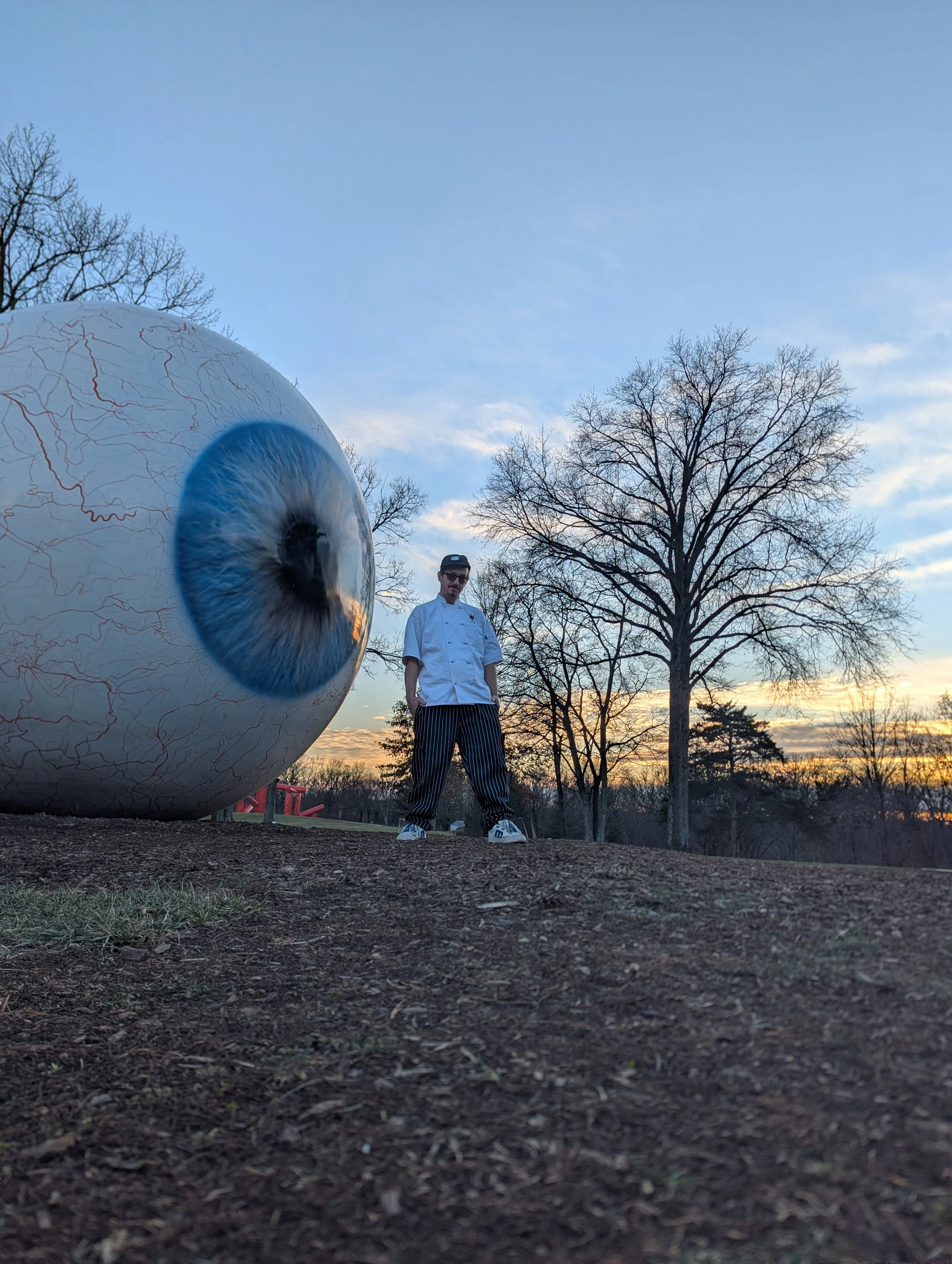 A person dressed as a chef standing outdoors next to a giant eyeball sculpture under a clear evening sky with trees and a sunset in the background.