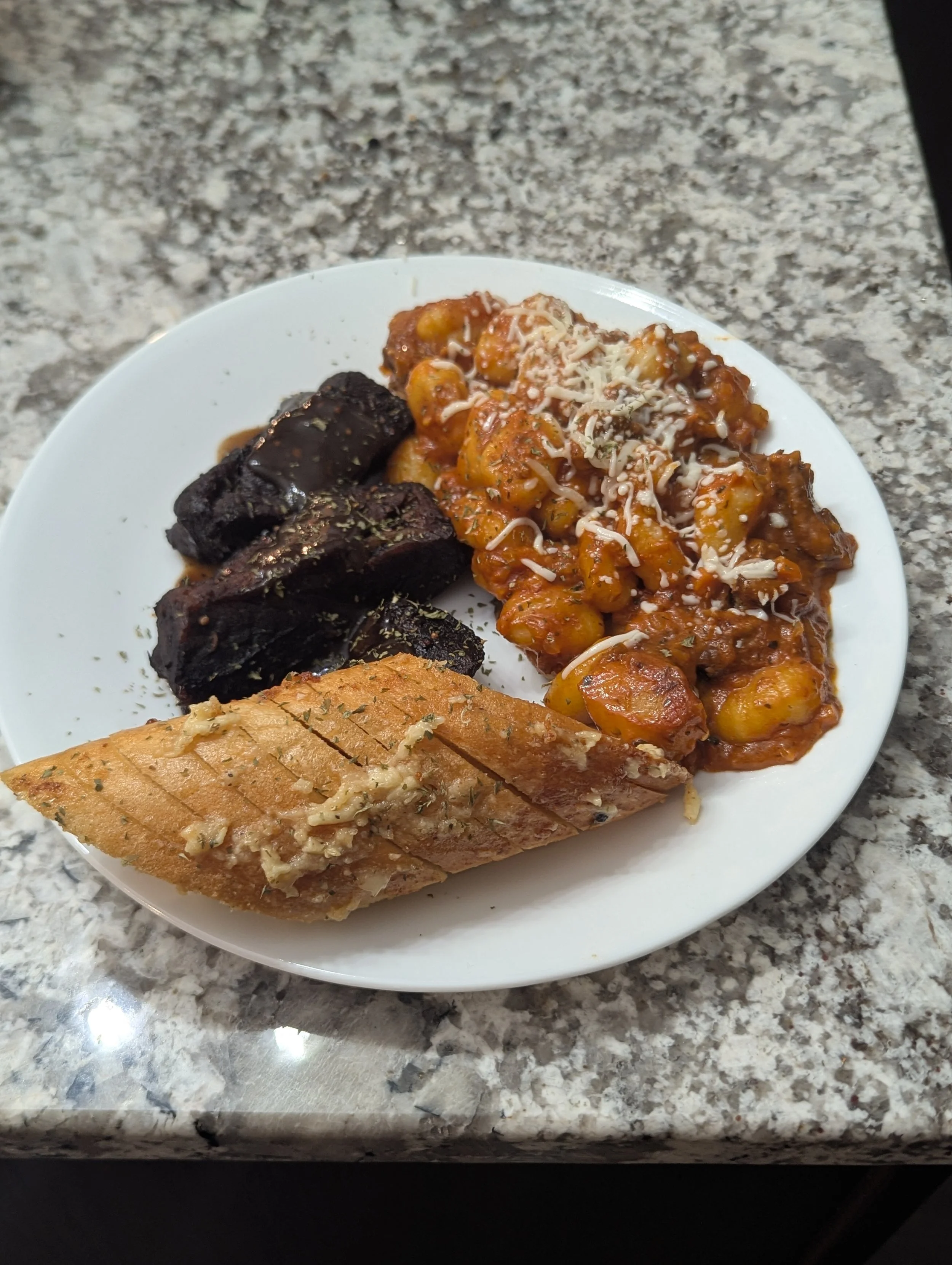 A white plate with grilled garlic bread, cooked chickpeas in tomato sauce topped with cheese, and slices of cooked beef on a granite countertop.