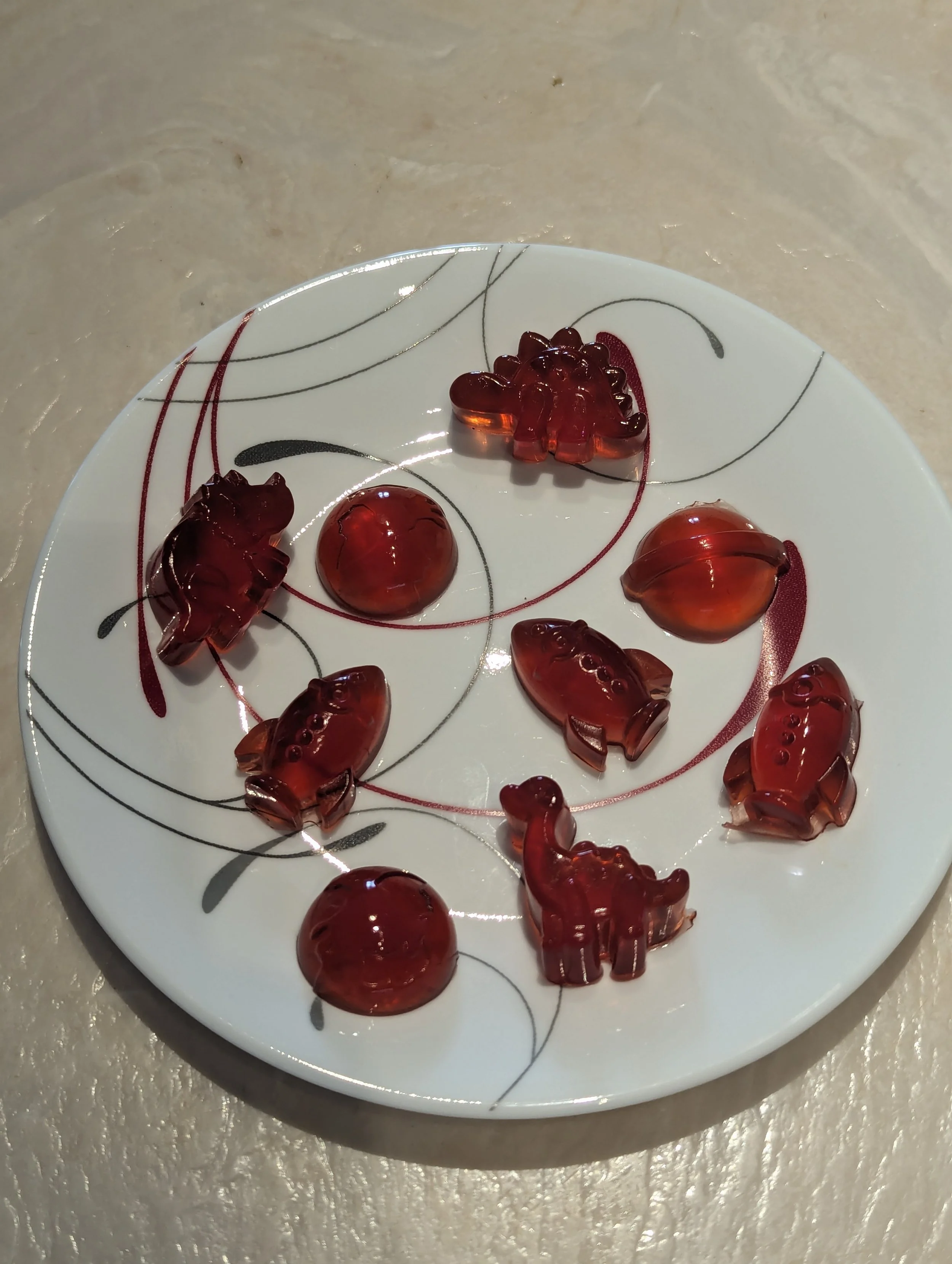 A white plate with black and red swirl patterns holding several small, cut-out gummy candies in various shapes, including animals and round shapes, made of translucent red gummy material.