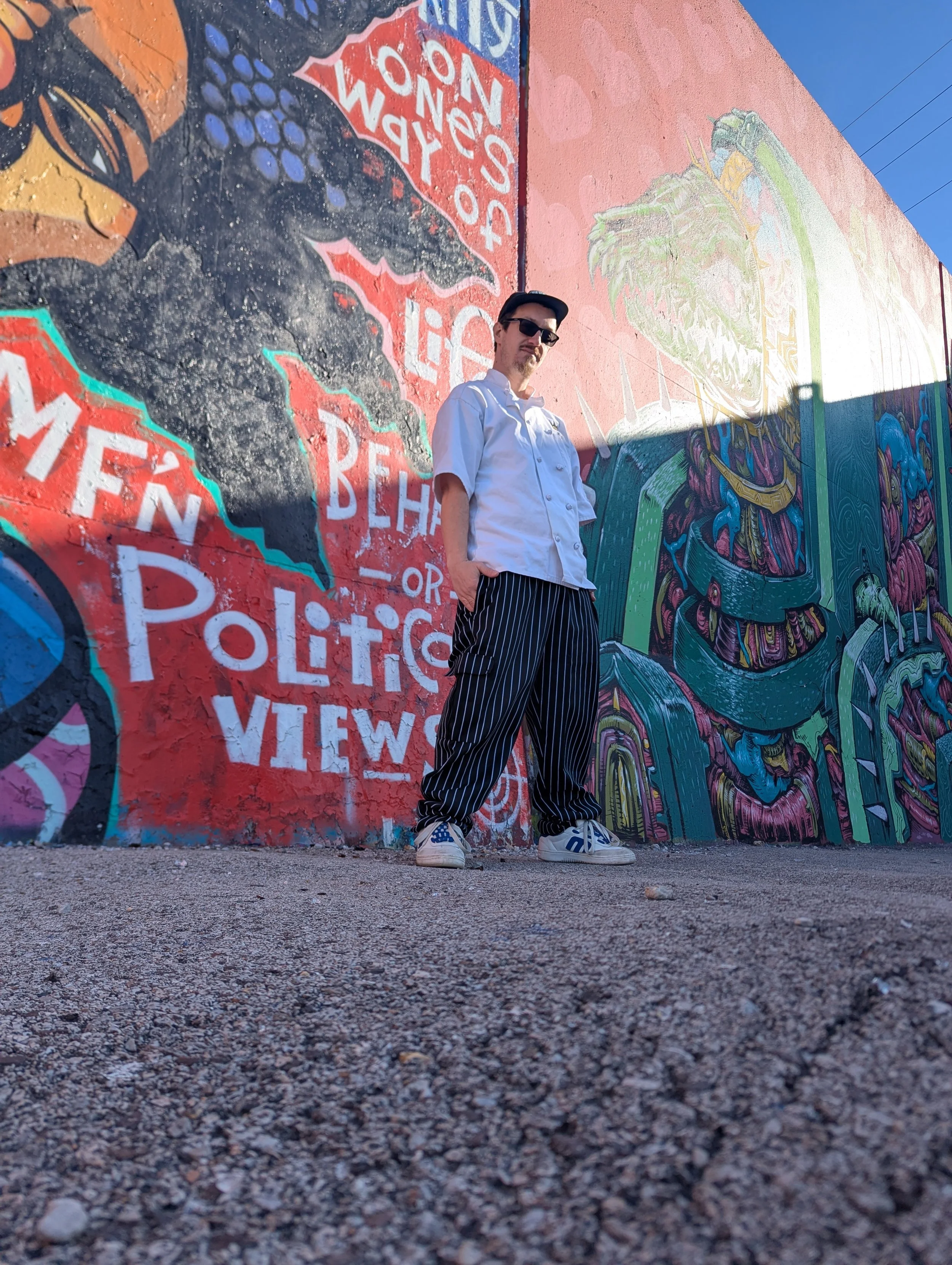 A person wearing sunglasses, a white chef's coat, and striped pants standing in front of a colorful mural with graffiti-style art and text such as 'Fear' and 'Politics Views'.