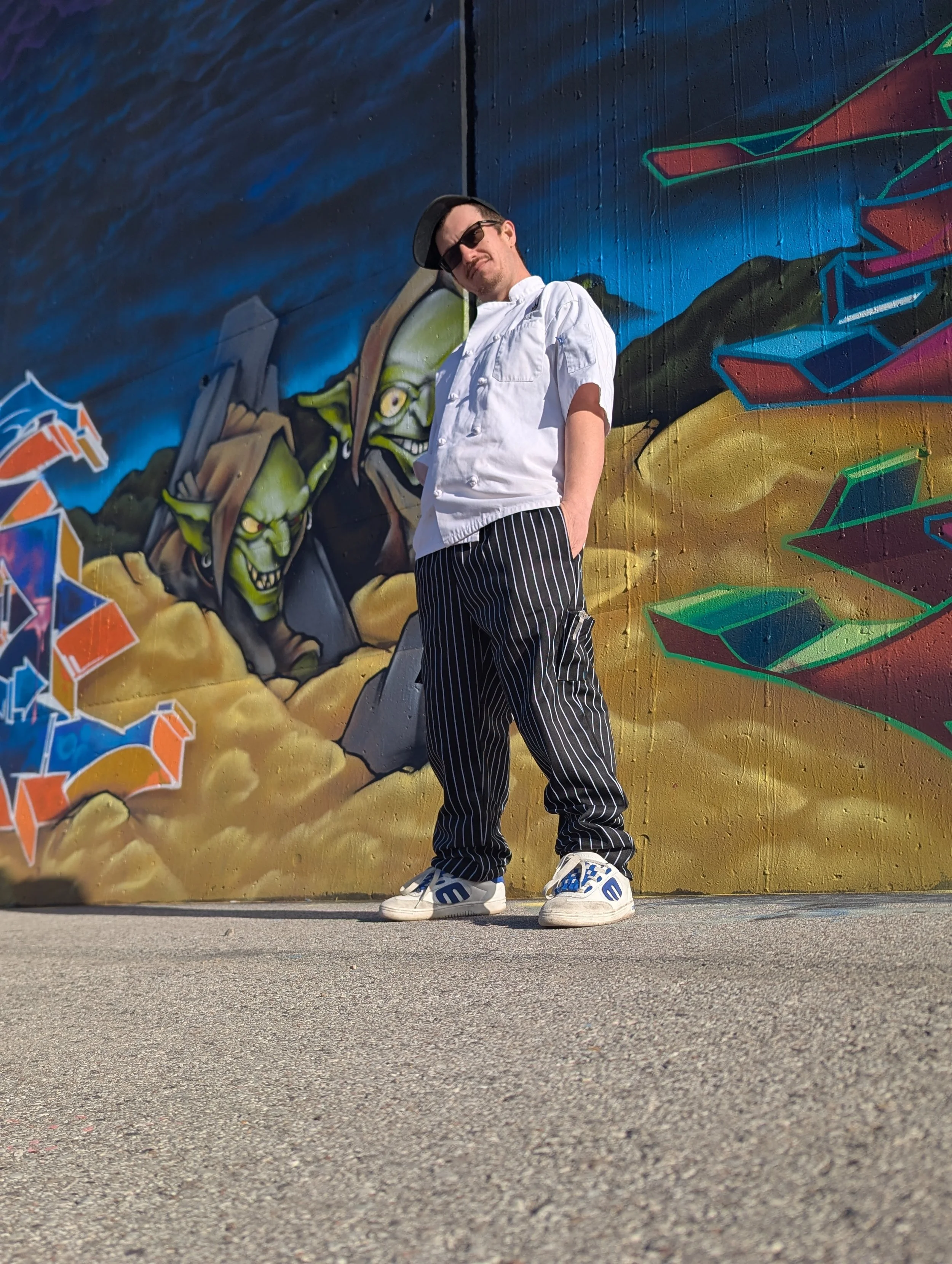 Man in white chef's coat, striped pants, and sneakers standing in front of colorful graffiti mural with goblin-like creatures.