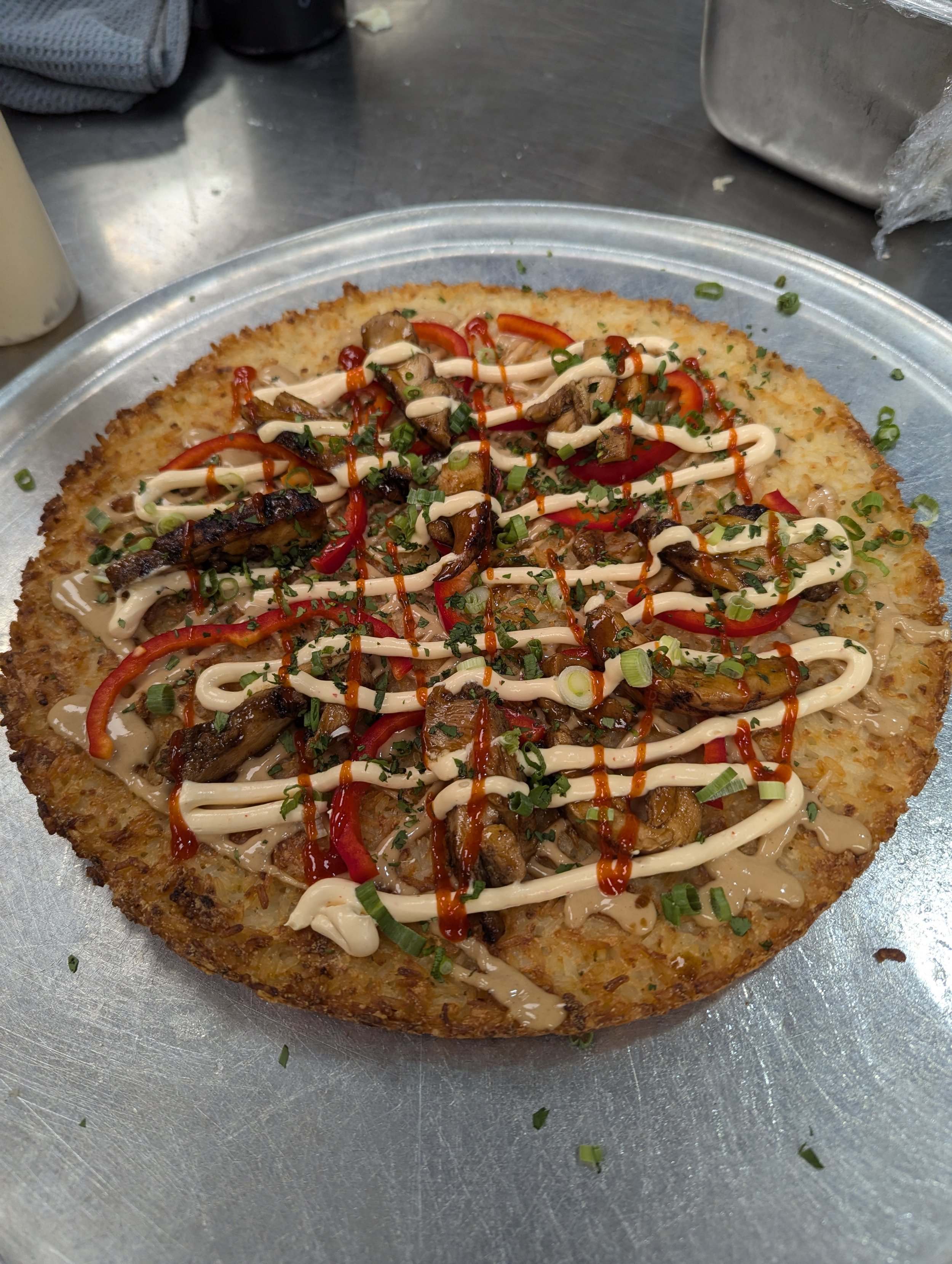 A savory Korean pancake topped with sautéed mushrooms, sliced red bell peppers, chopped green onions, mayonnaise, and hot sauce on a metal plate.