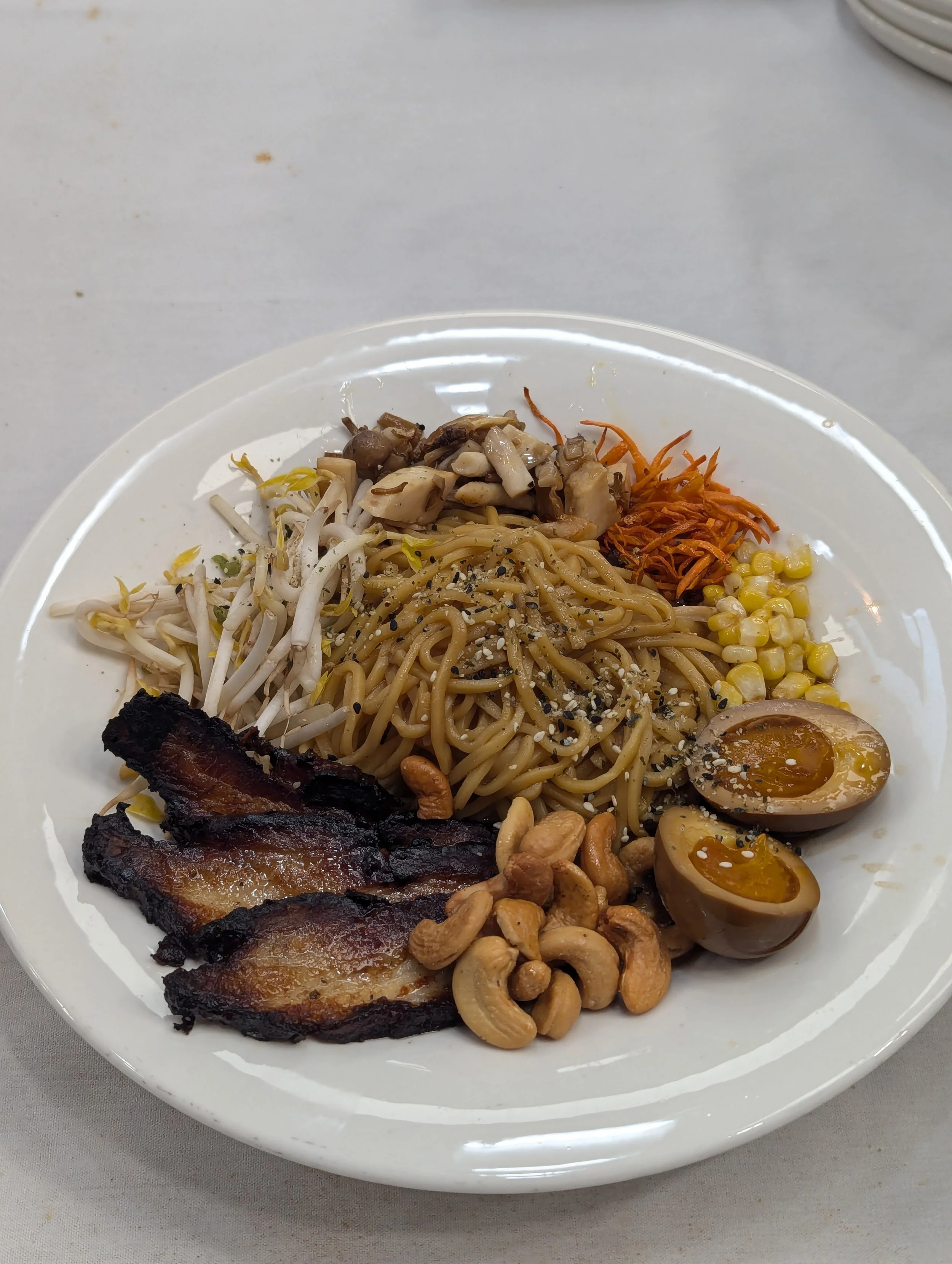 Asian-inspired noodle bowl with grilled pork belly, soft-boiled eggs, bean sprouts, shredded carrots, corn, chopped mushrooms, and cashews.