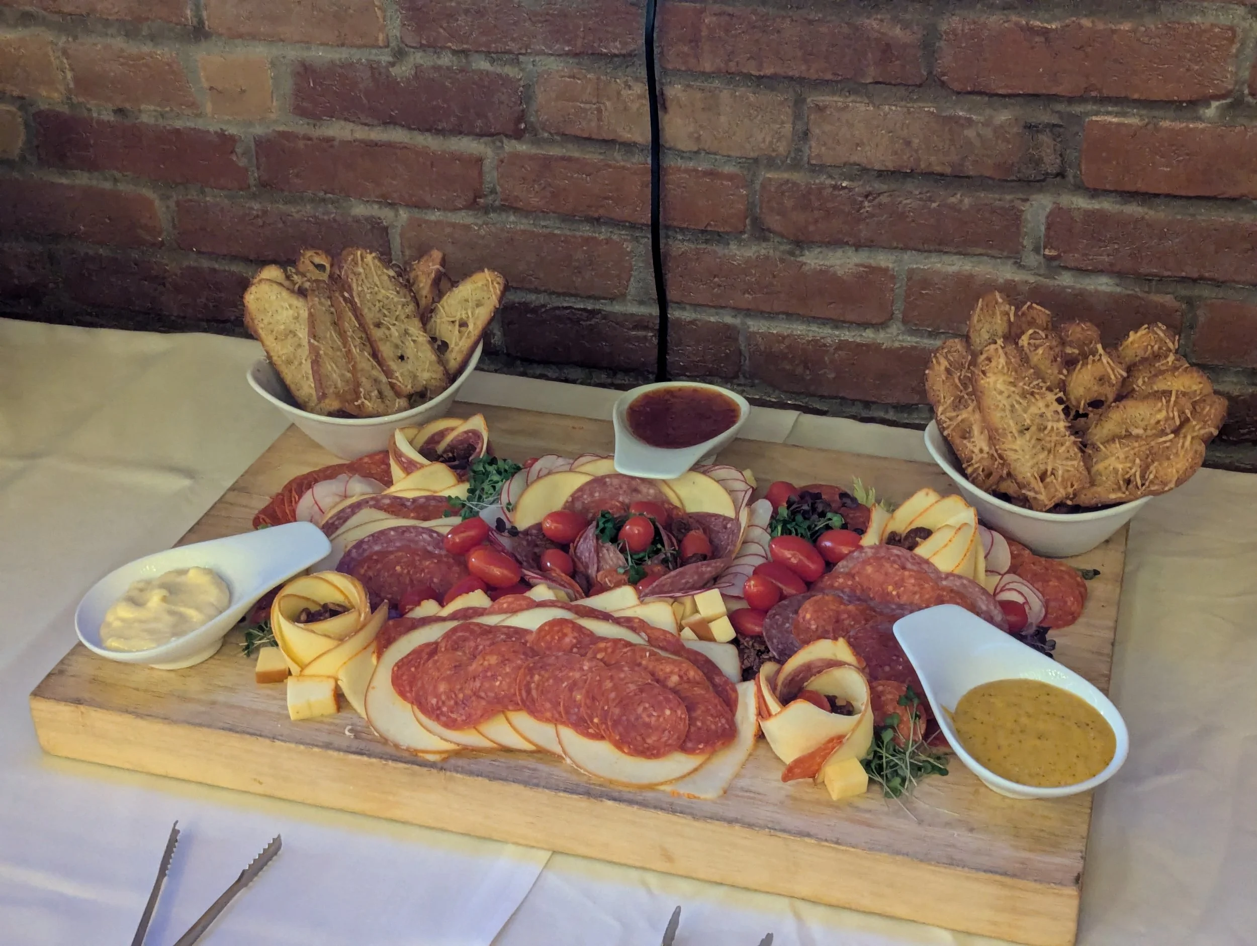 A charcuterie board with assorted cured meats, cheeses, cherry tomatoes, and garnishes, accompanied by bowls of fried plantains, dipping sauces, and a small bowl of honey mustard, set on a wooden board against a brick wall.