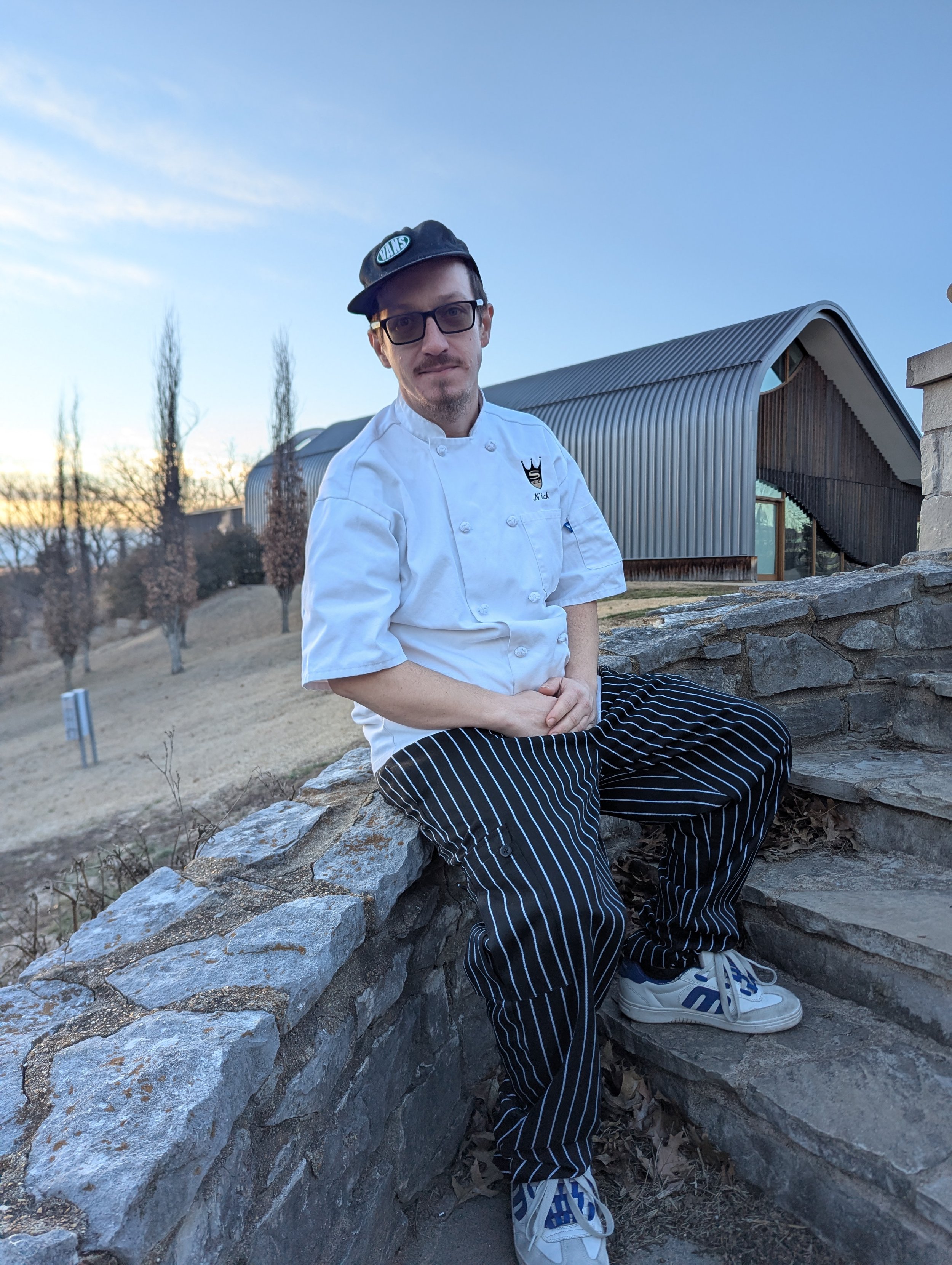 A man dressed as a chef sitting outdoors on a stone wall with a modern building in the background, wearing a white chef's coat, striped pants, and a black cap.