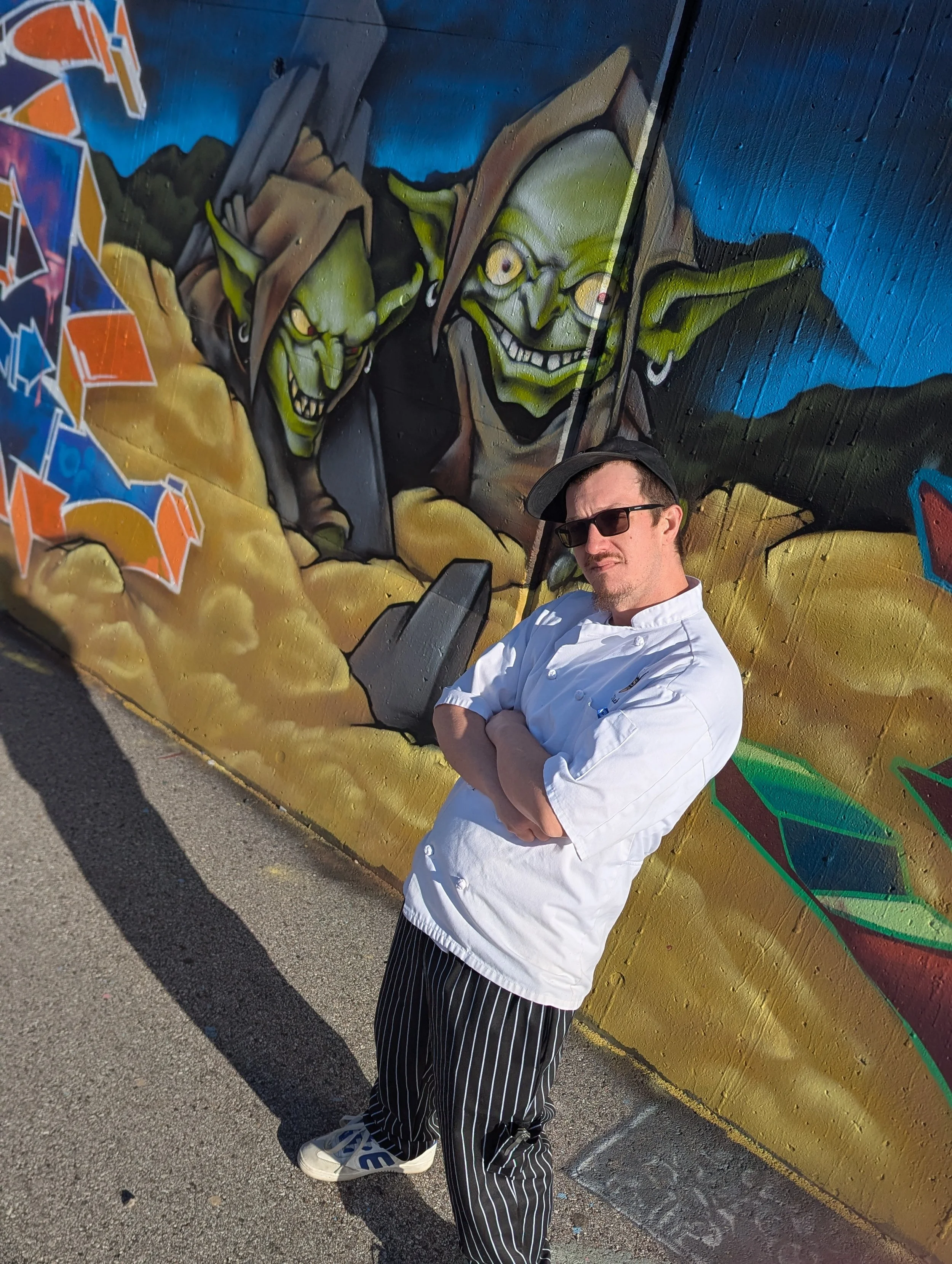 A man dressed as a chef with crossed arms and sunglasses standing in front of a colorful graffiti mural featuring two goblin characters.