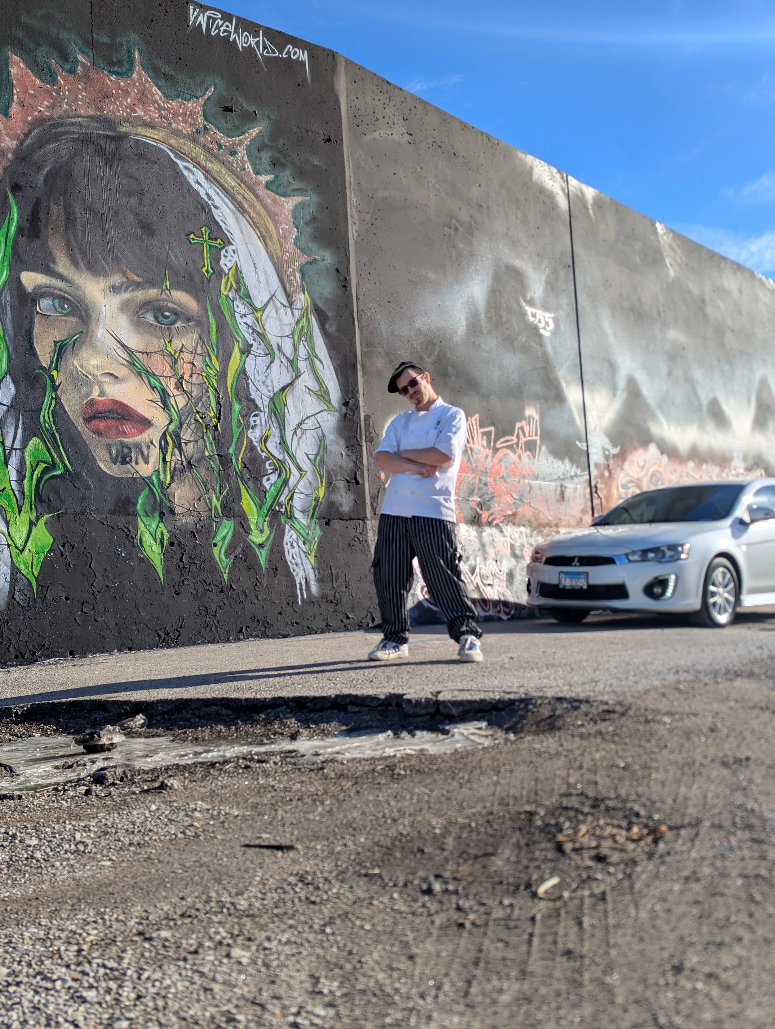 A man standing with arms crossed in front of a large mural of a woman’s face with green vines and crosses painted on her face, on a city street during daylight. There is a silver car parked nearby.