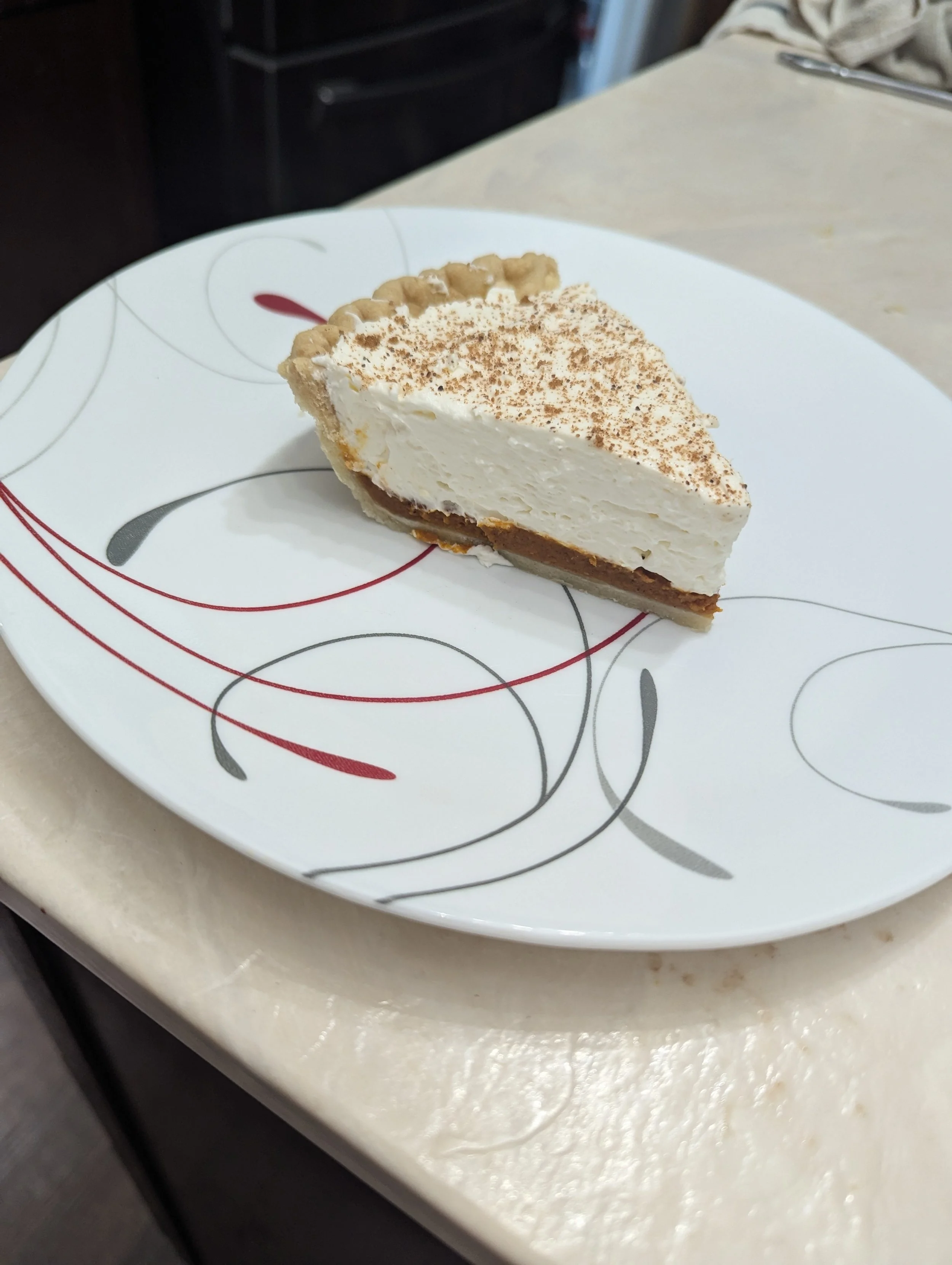 A slice of cheesecake with a graham cracker crust, topped with whipped cream, sprinkled with cocoa powder, on a white plate with decorative red and gray swirls, placed on a light-colored table.