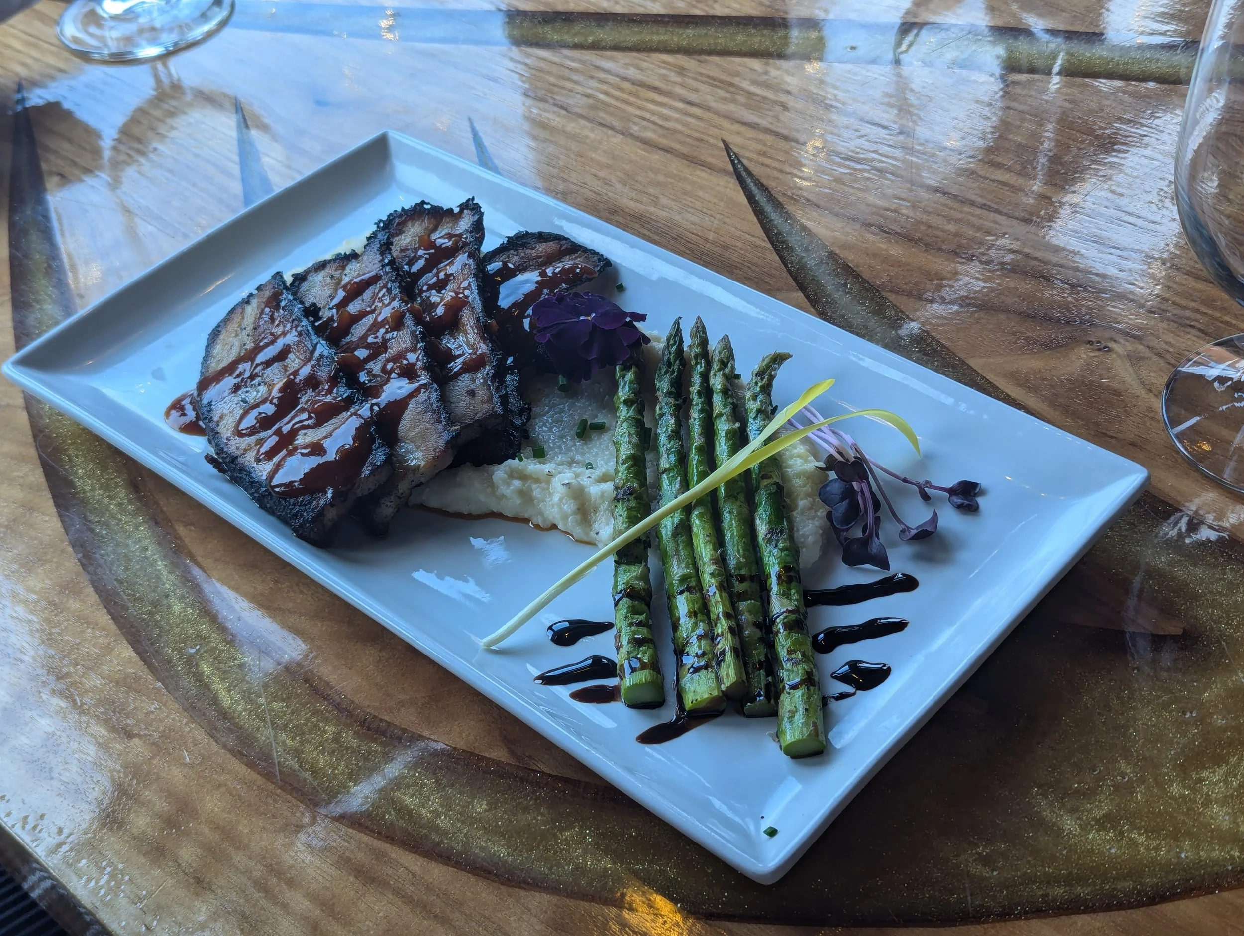 A rectangular white plate with sliced grilled meat topped with barbecue sauce, grilled asparagus spears drizzled with balsamic glaze, cauliflower mash garnished with edible flowers and microgreens, all on a round wooden table with a glass of water an
