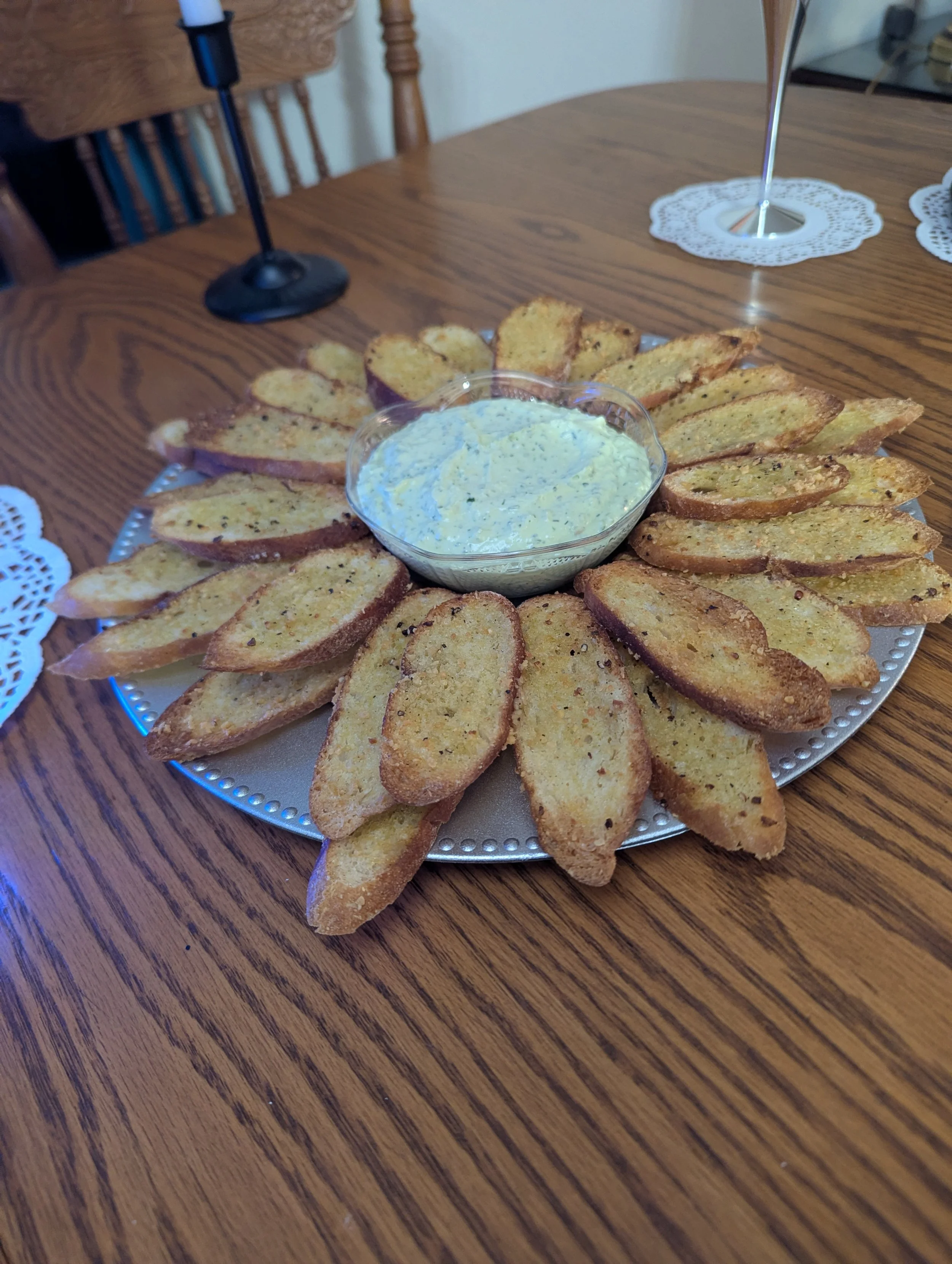 A round platter of toasted garlic bread slices arranged around a bowl of dipping sauce on a wooden table.