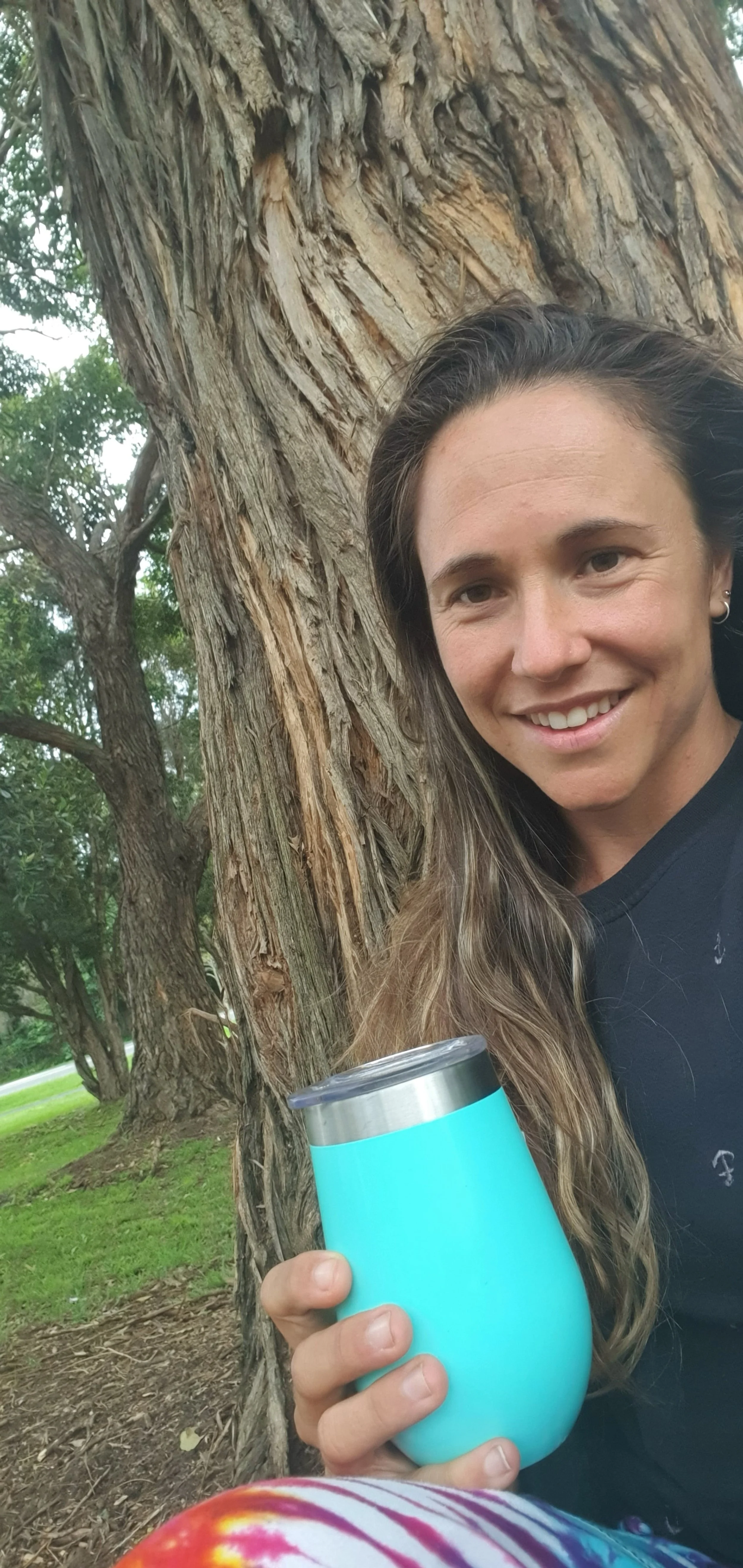 A woman with long hair holding a light blue insulated tumbler, standing next to a large tree in a park.