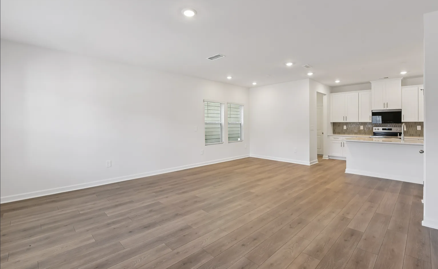 Empty living room with white walls, hardwood flooring, two windows with blinds, and a kitchen in the background featuring white cabinets, a granite countertop, and stainless steel appliances.