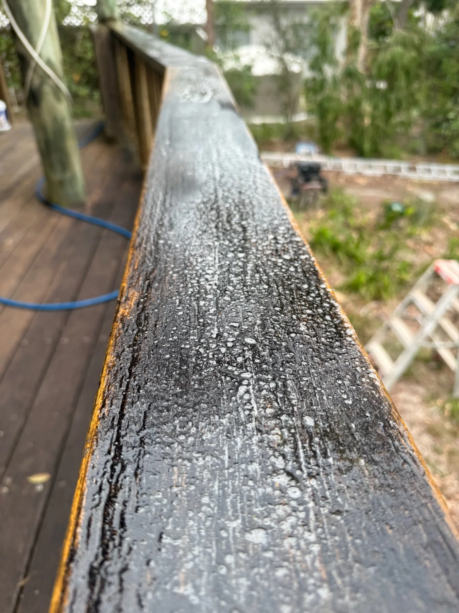 Close-up of a wet, black wooden railing with water droplets on it, part of an outdoor deck with a blurred background of trees and other outdoor structures.