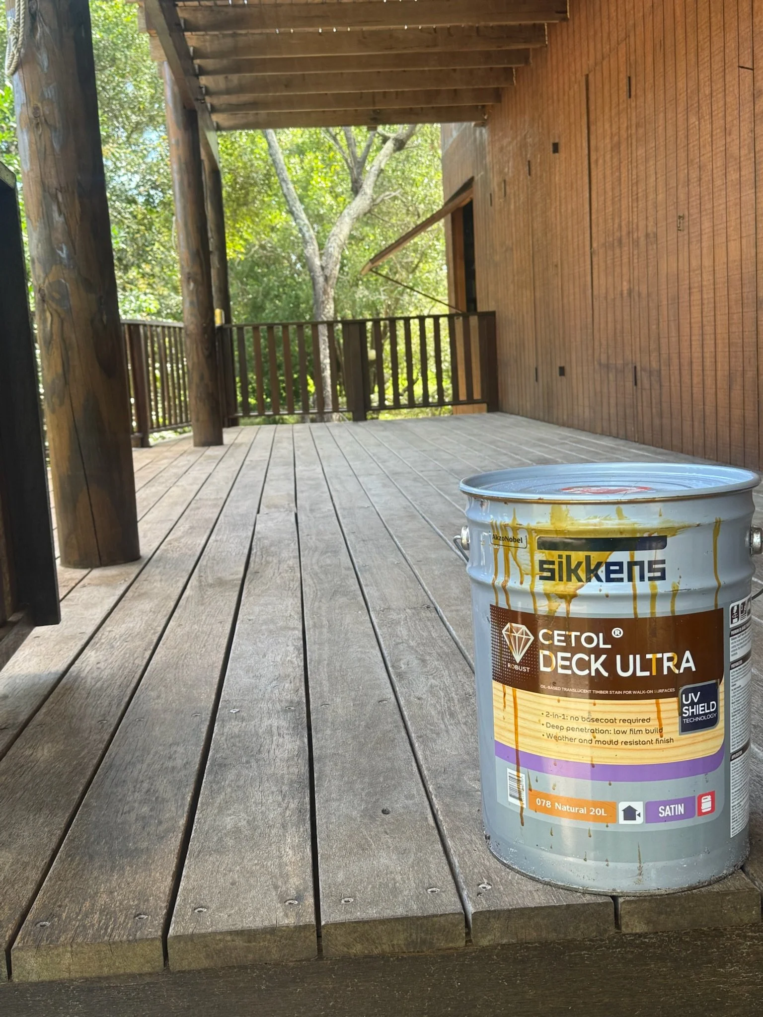 A wooden deck on an outdoor porch with a can of Cetol Deck Ultra stain placed on it, surrounded by a wooden railing and trees in the background.
