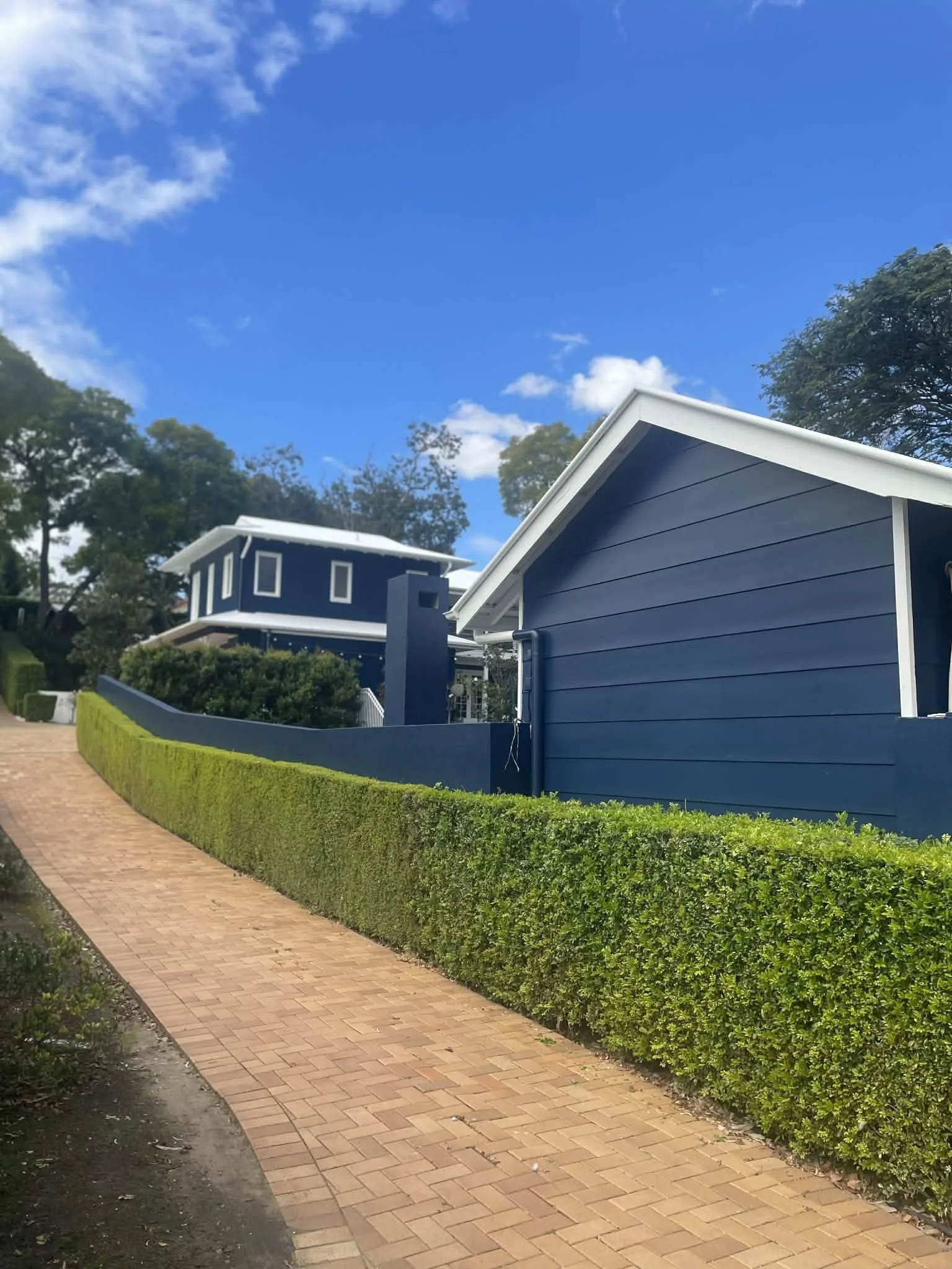 A sidewalk runs alongside a well-manicured hedge in front of blue-colored houses with white trim and slanted roofs, under a partly cloudy blue sky.