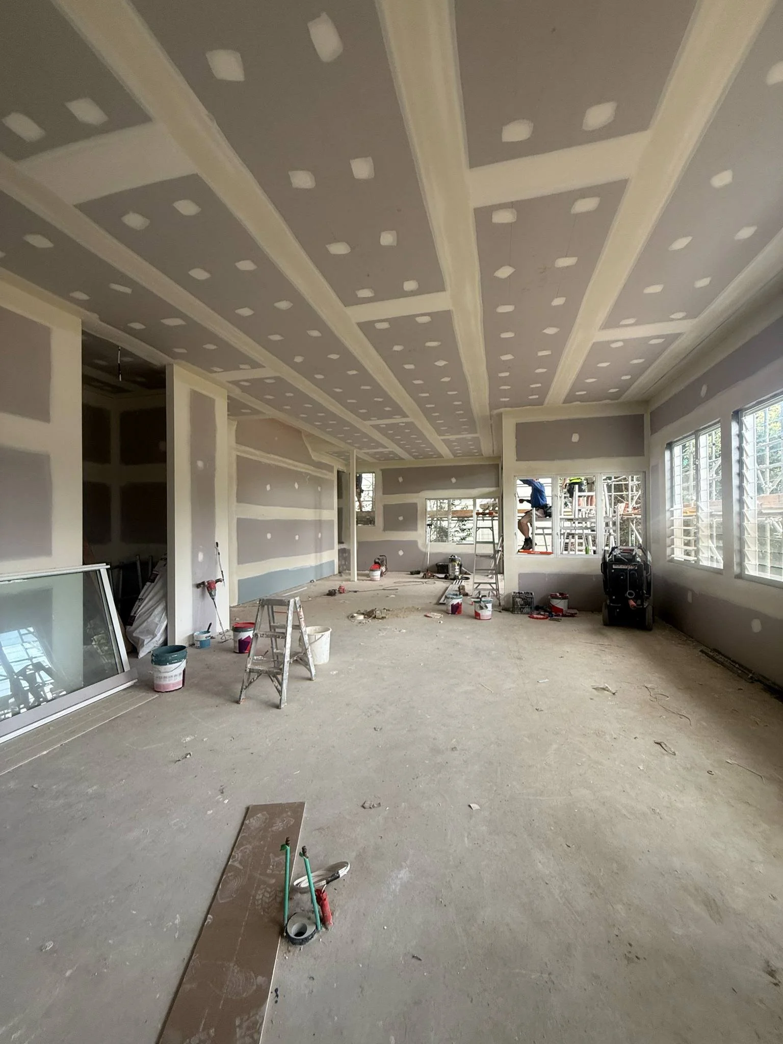 An unfinished room under construction with drywall installed on walls and ceiling, with visible joints and screws, and construction tools and supplies scattered on the floor, including a ladder, buckets, and a worker on a scaffold outside the window.