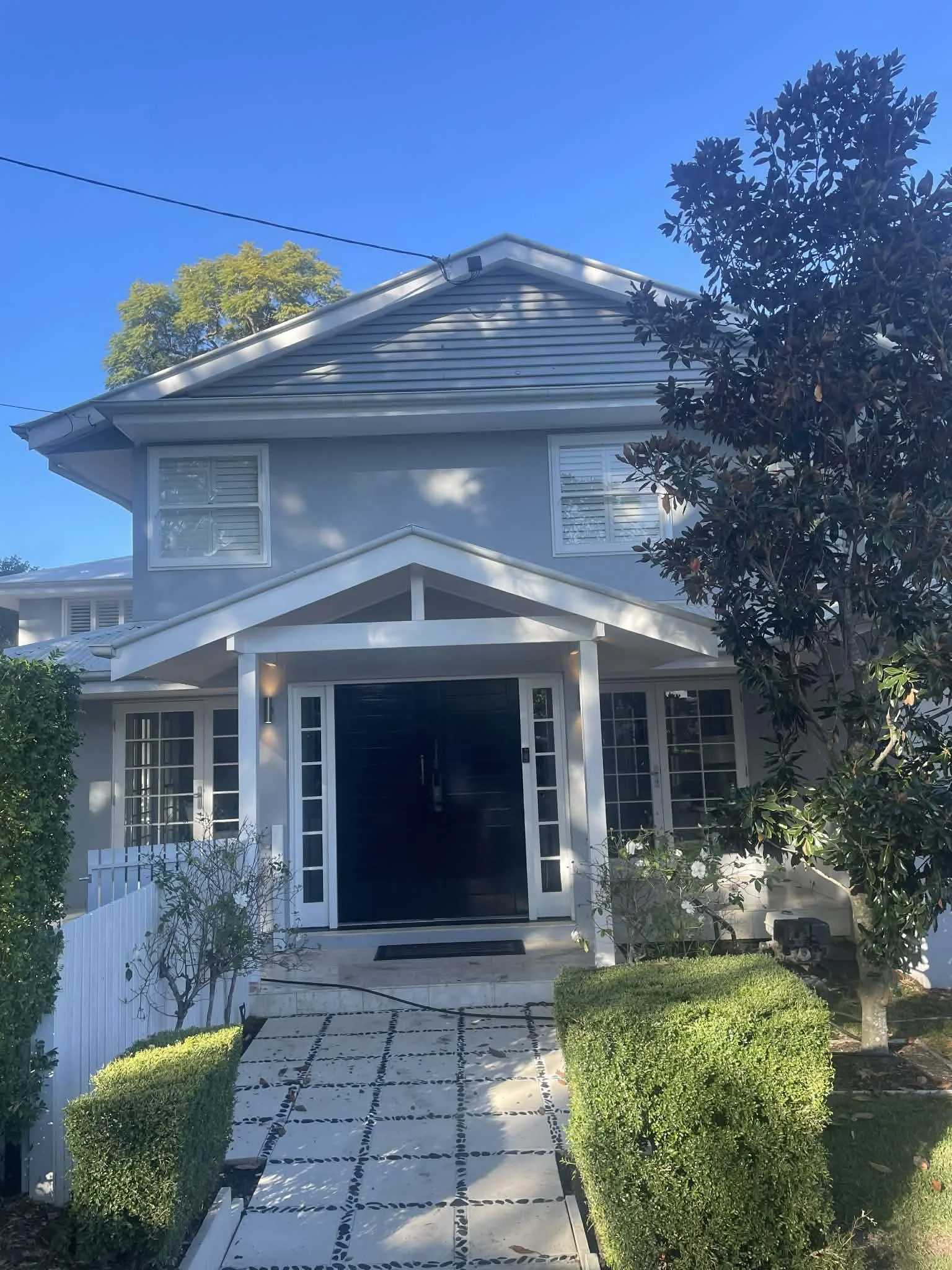 Front view of a two-story house with a black front door, white trim, and large windows, surrounded by a manicured lawn and trees under a clear blue sky.