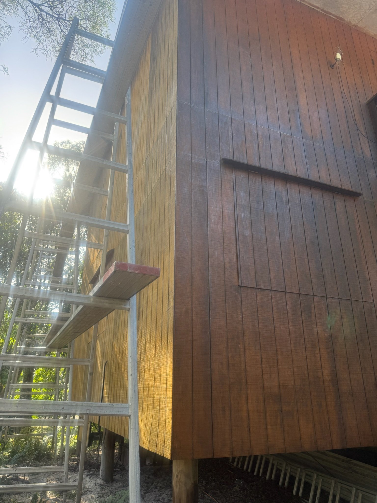 Construction site with wooden deck and metal ladder, sunlight shining through trees, and a small outdoor light fixture on beige wall.